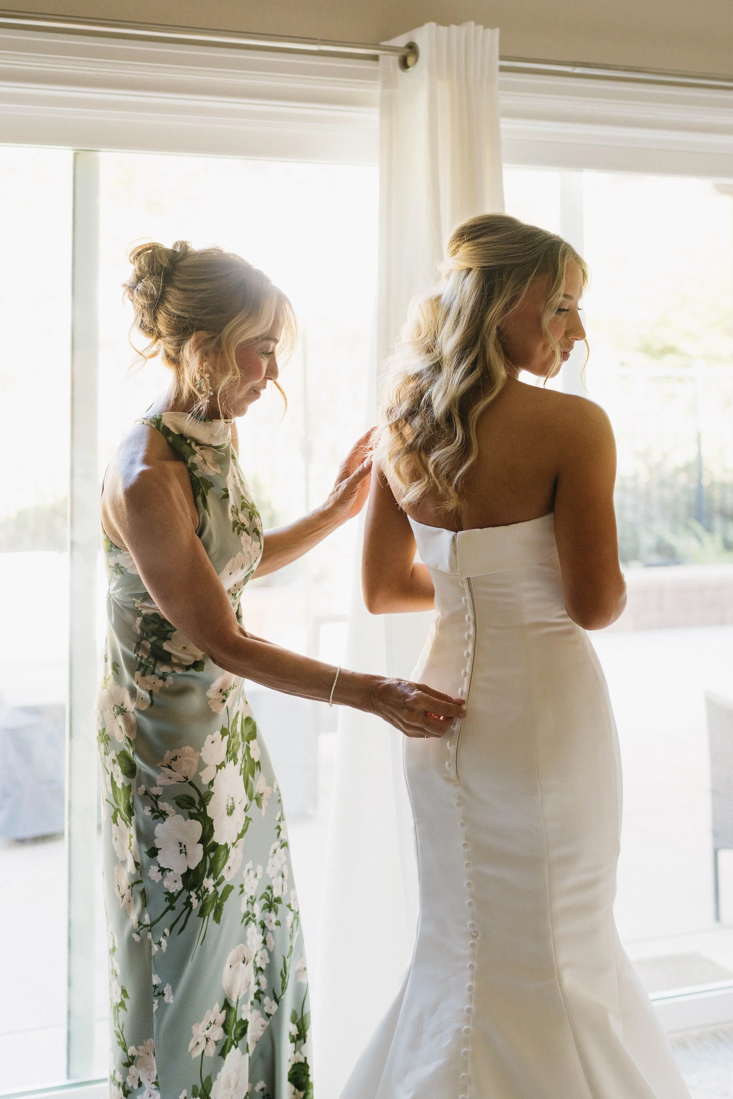 A bride with wavy hair half-up half down in a strapless white wedding gown is being helped with her dress by her mother who has a curly updo wearing a green floral dress, near a large window with sheer white curtains.