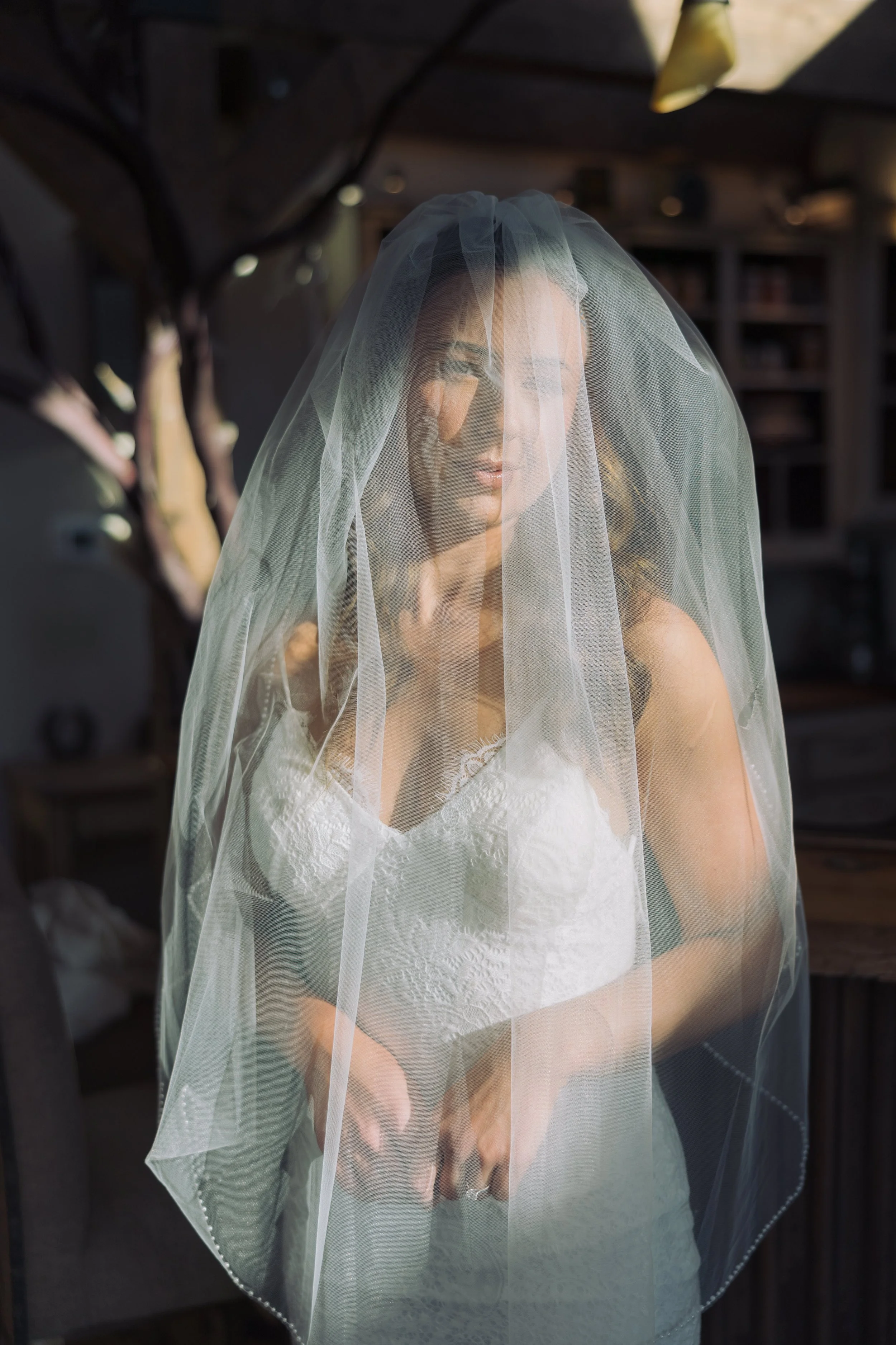 A bride wearing a white lace top and a veil, standing indoors with bookshelves in the background as natural light comes in.