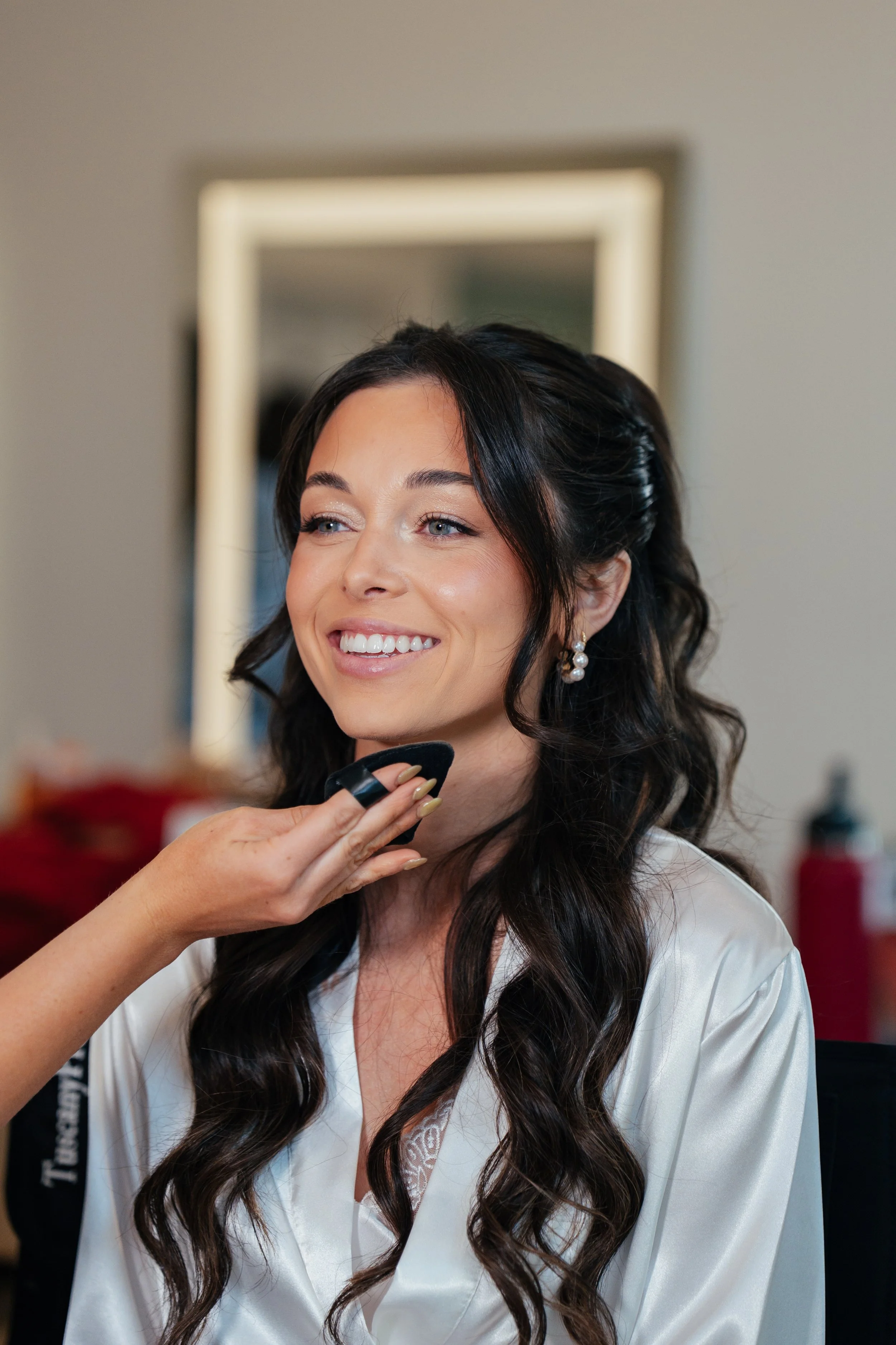 A bride with long, dark, wavy hair in a white satin robe smiling as someone powders the bottom of her chin.