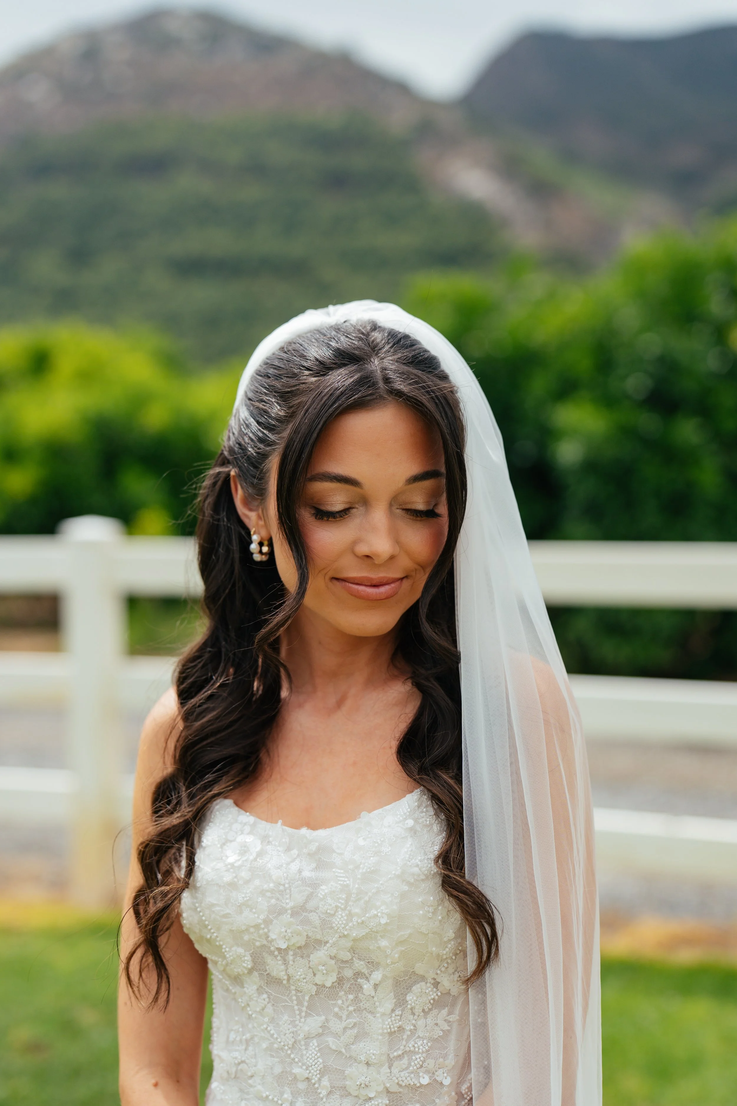 Close-up of a bride with long dark wavy hair, natural soft makeup, a white lace wedding dress and veil with floral embellishments, outdoors with mountains and green foliage in the background.
