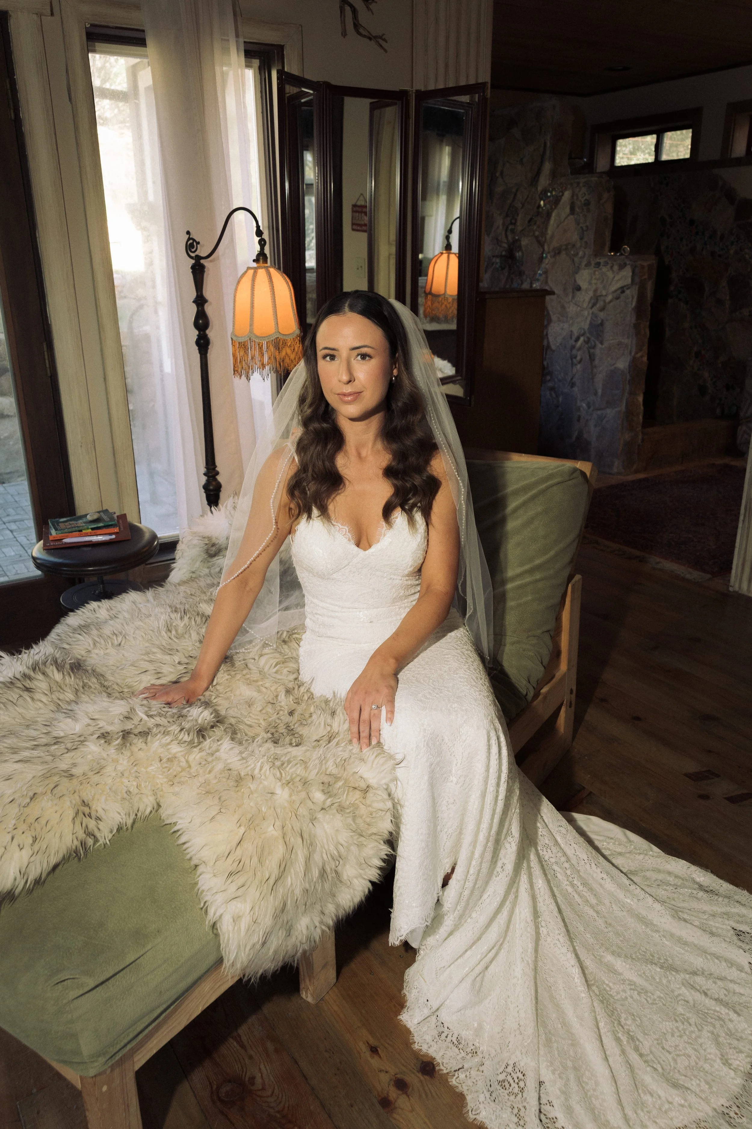 A bride sitting on a couch wearing a white wedding dress and veil, with long brown hair and natural glam in a cozy room with wooden walls and stone fireplace, next to a window with open curtains and a floor lamp.