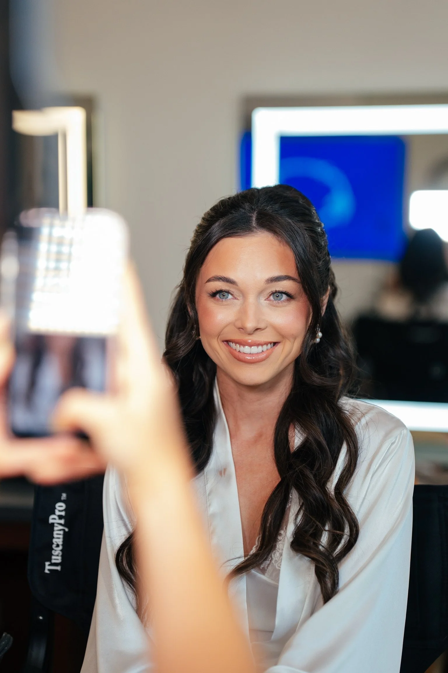 A woman with long dark hair and blue eyes smiling while someone takes her picture with a smartphone.