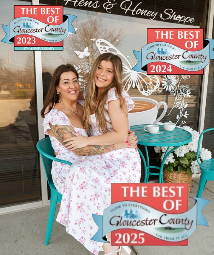Two women sitting together outside a cafe, smiling and hugging. They are wearing matching white dresses with pink floral patterns. There are stickers on the image indicating awards for 'The Best of Gloucester County' for the years 2023, 2024, and 2025. The background shows a large coffee mug graphic, a potted plant, a white butterfly, and some white floral decorations.