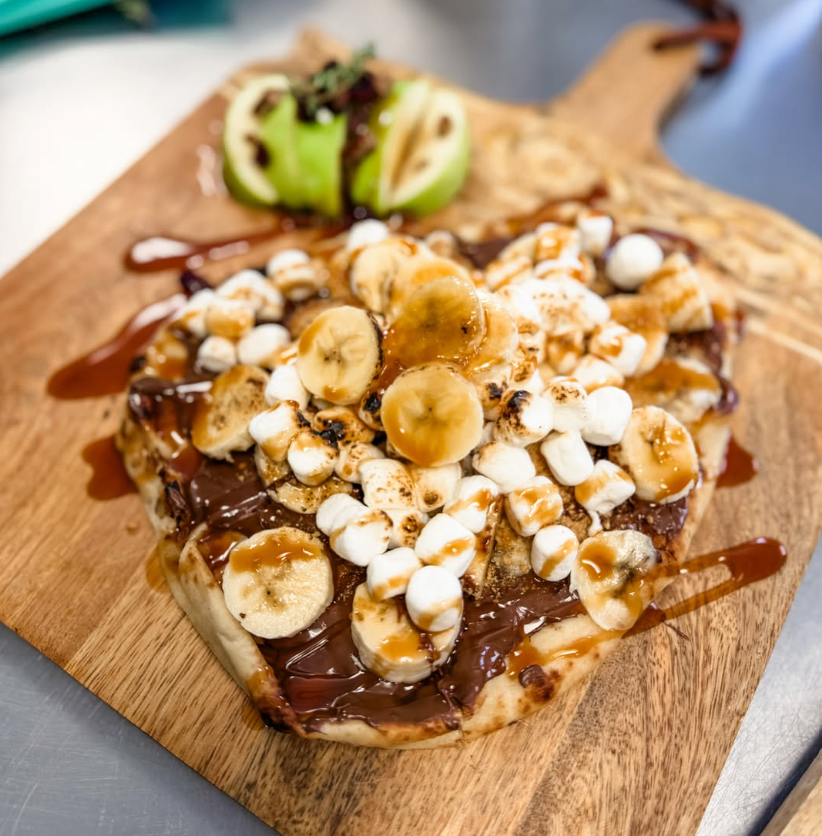 Chocolate banana s'mores pizza with marshmallows, banana slices, and drizzled caramel on a wooden serving board.