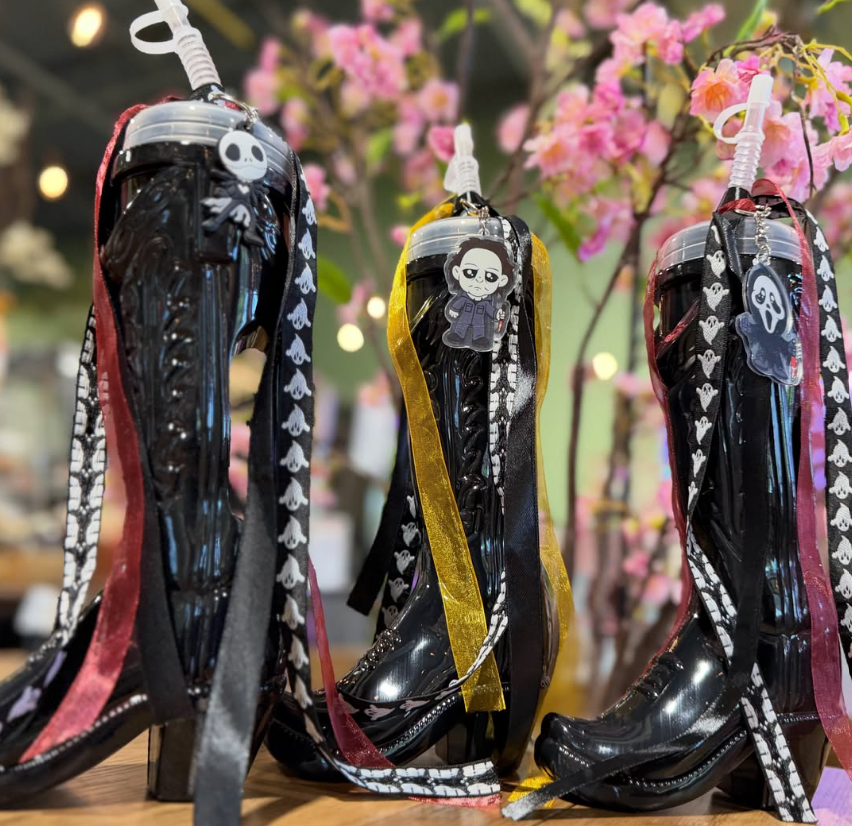 Decorative glass high-heeled boots with ribbons and Halloween-themed charms, displayed in front of pink flowers.