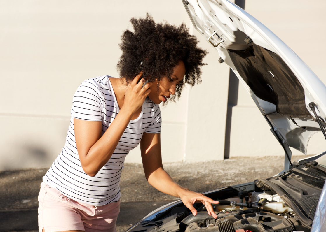 A young woman with curly hair in a striped shirt and pink shorts looking worried while checking her car engine with the hood open.