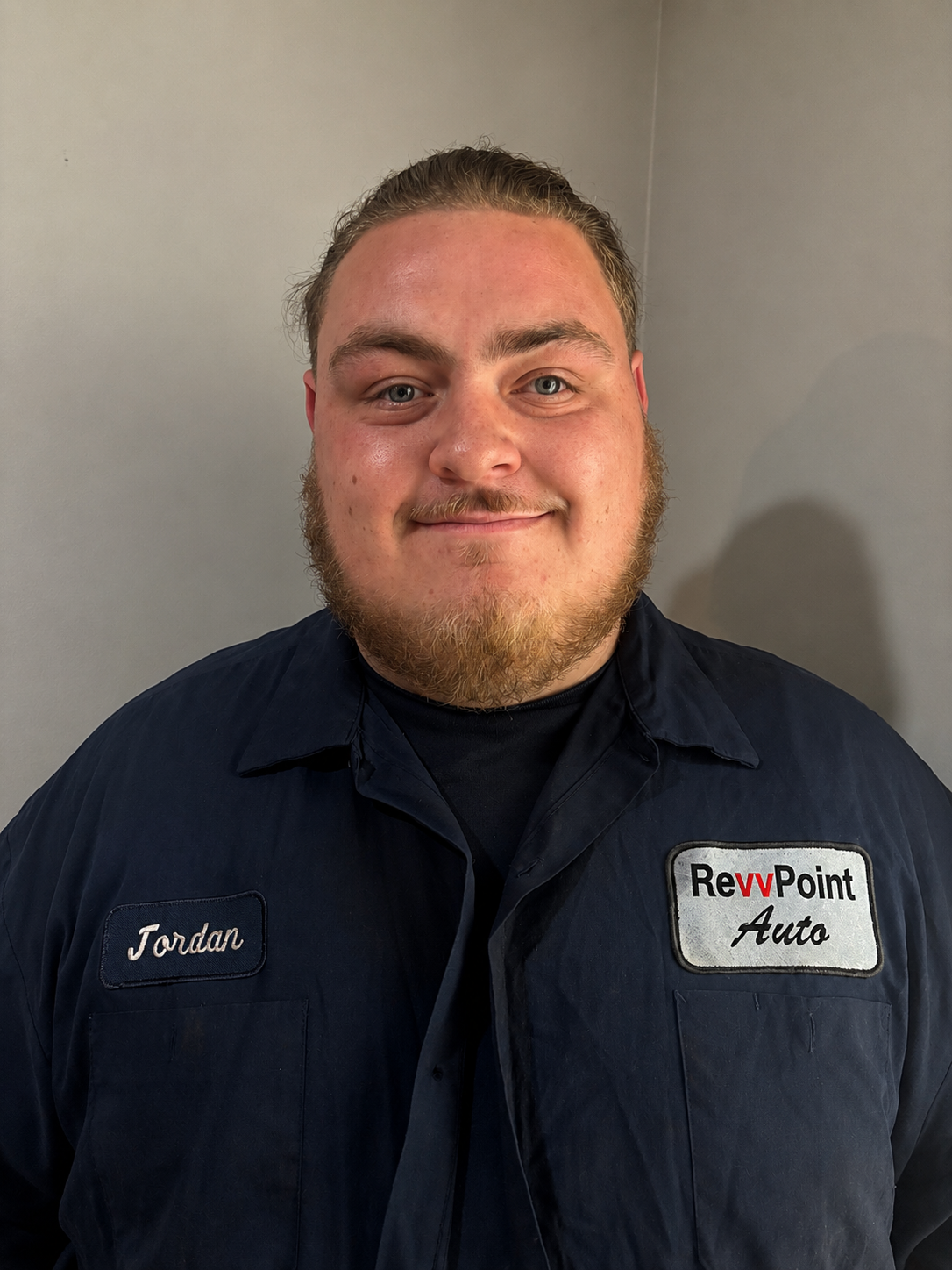 A man with a beard and mustache wearing a navy blue work uniform with the name 'Jordan' on it and an 'RevvPoint Auto' patch, standing against a plain wall.