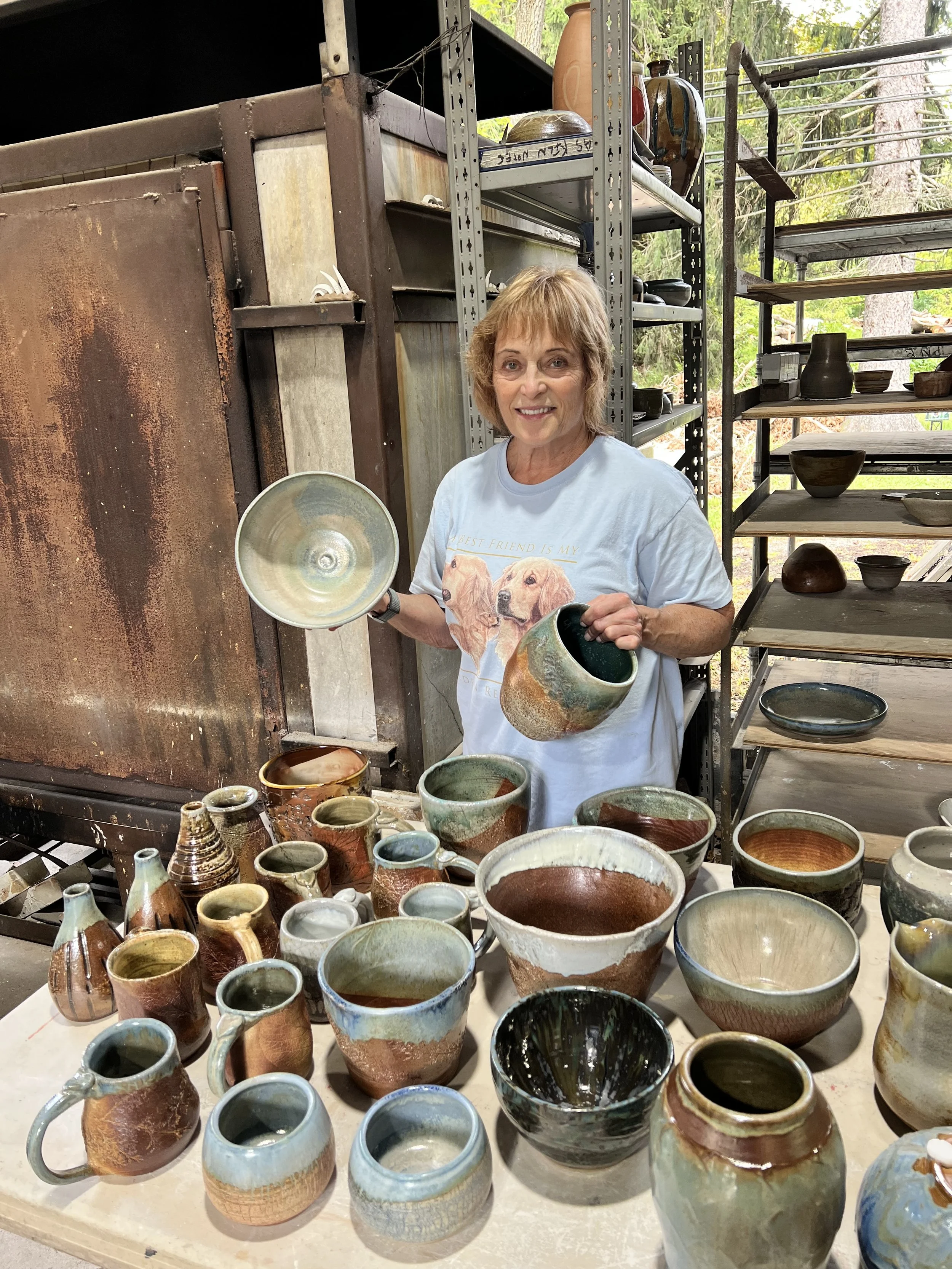 A smiling woman holding pottery mugs in a workshop filled with various handmade pottery items on tables and shelves, with a background of trees and outdoor scenery.