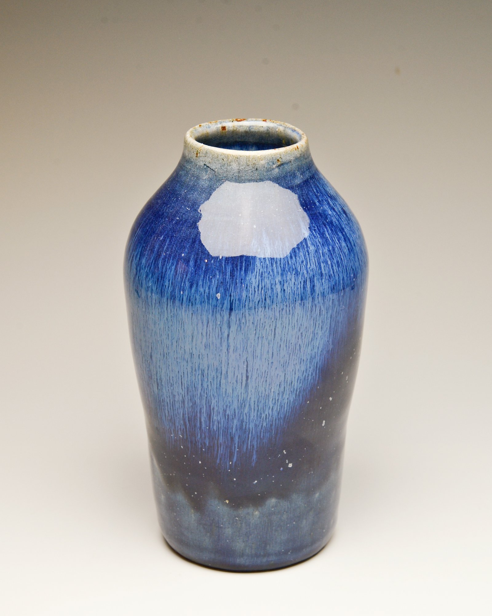 A ceramic vase painted in a gradient of blue hues, from light to dark, with a rough, textured surface and a narrow opening at the top.