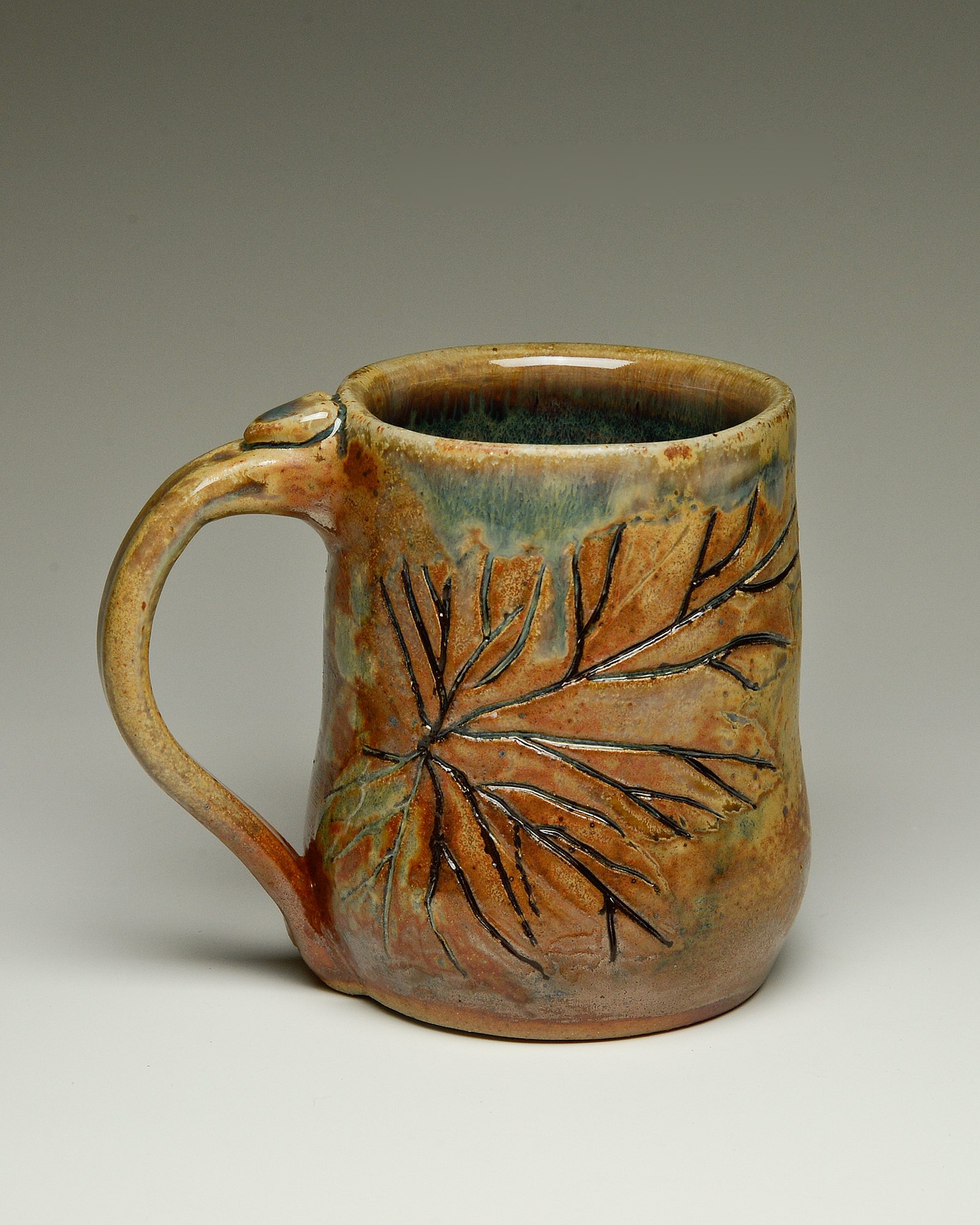 A ceramic mug with leaf designs, glazed in warm earth tones, featuring an outside depicting a detailed leaf pattern.