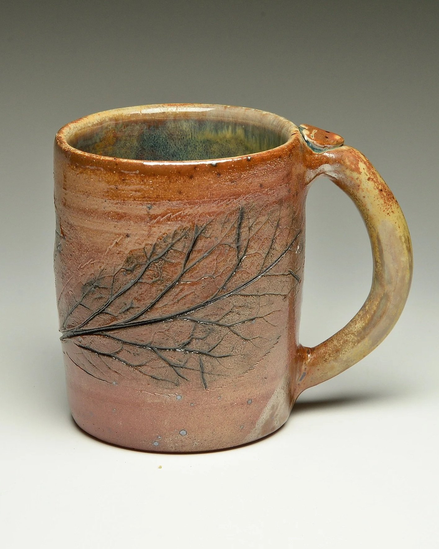 Ceramic mug with a leaf pattern engraved on its surface, glazed with earth tones including reddish, yellow, and green hues, and a curved handle.