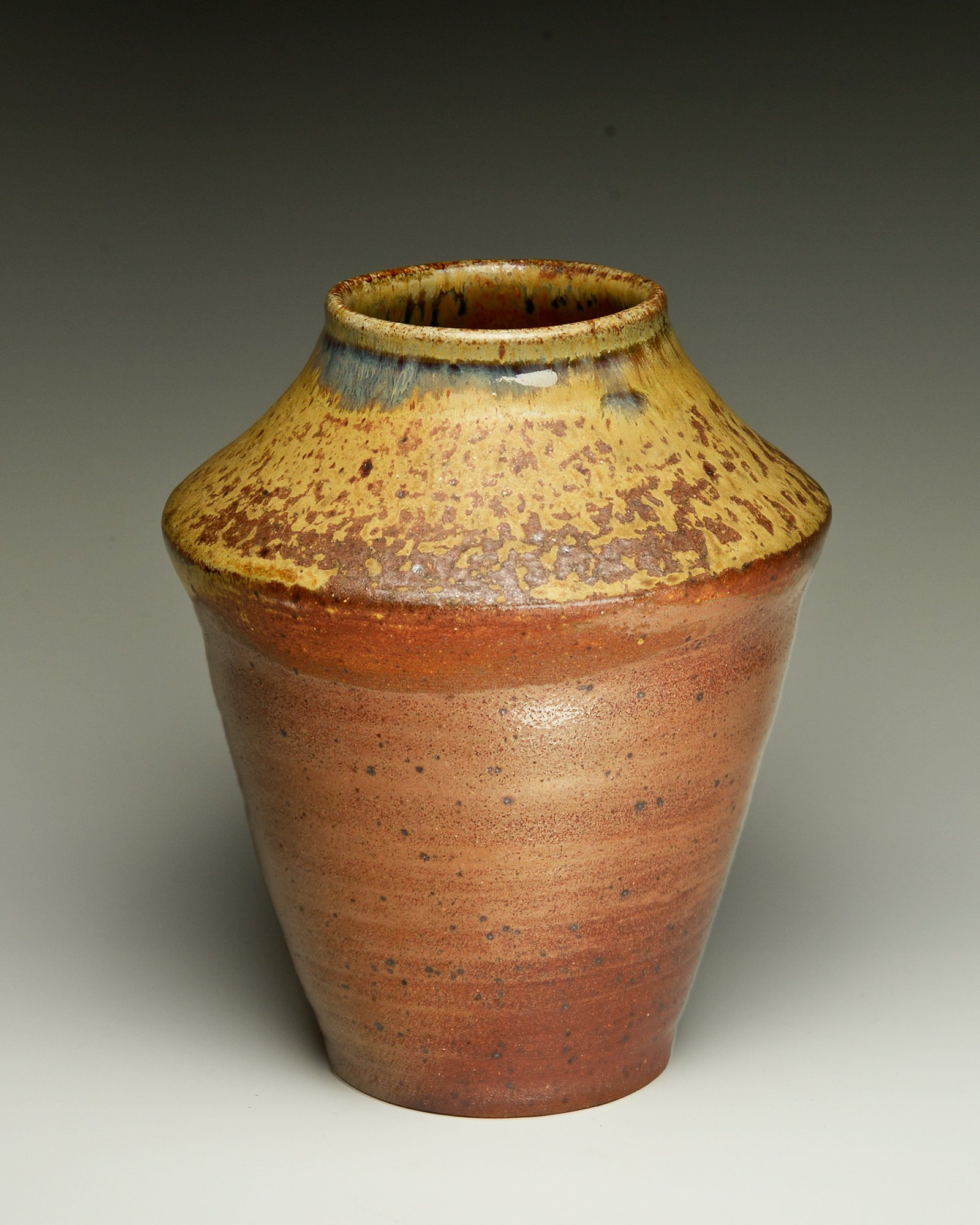 Close-up of a handcrafted ceramic vase with a rounded top, glazed in earthy tones of yellow, orange, and brown, featuring speckled and textured glaze patterns.