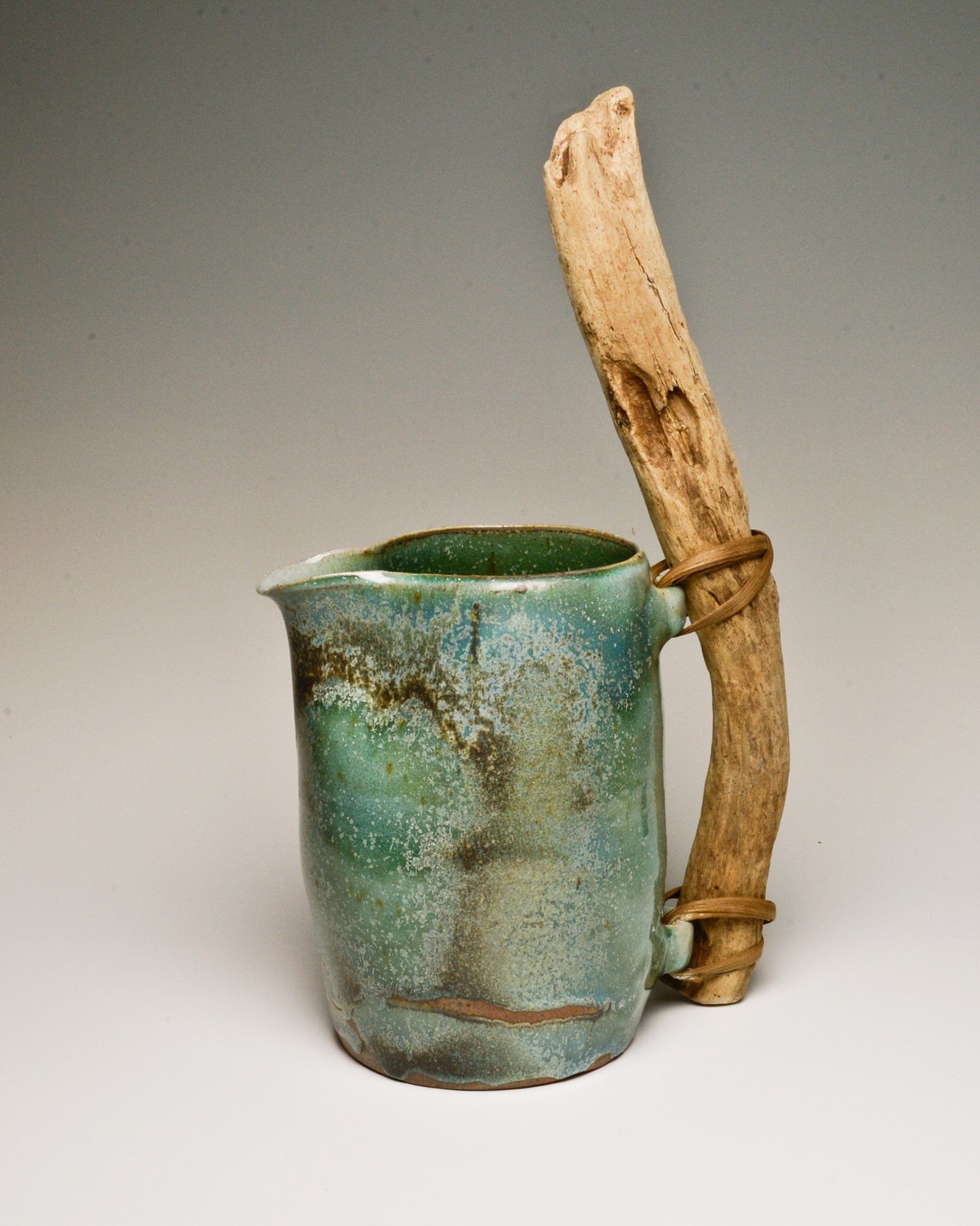 An old ceramic tea cup with a rustic glaze and crackles, accompanied by a weathered piece of driftwood tied to the cup with a rubber band.