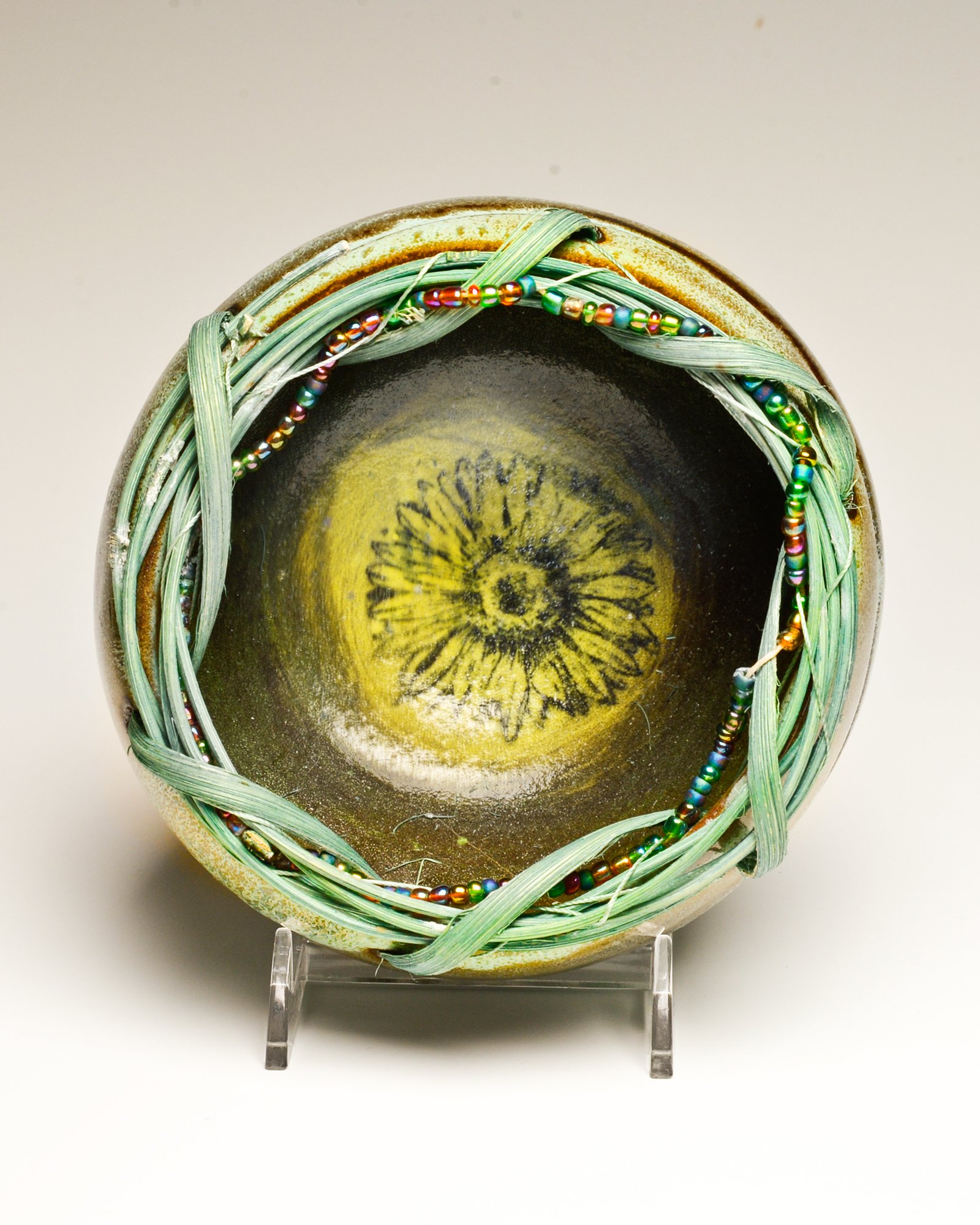 Top view of a ceramic vessel with a black interior, decorated with floral patterns, wrapped with strands of multicolored beads and green plant fibers, displayed on a clear stand.