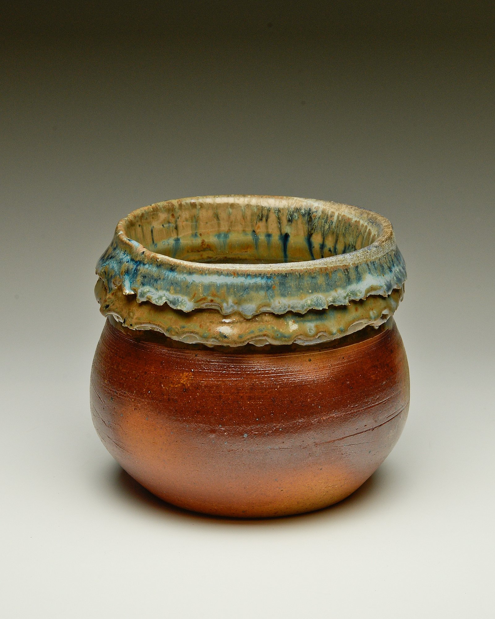 A ceramic pot with a reddish-brown base and a wide, decorative rim featuring a textured, dripping glaze in shades of blue, green, and beige.