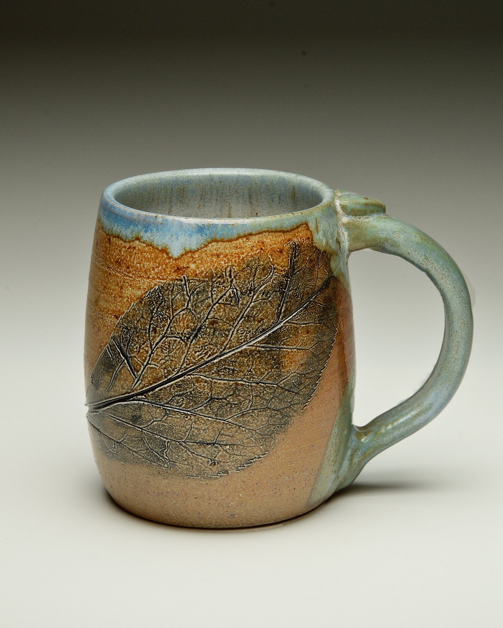 A ceramic mug with a textured leaf design and a color gradient from blue to brown, set against a neutral background.