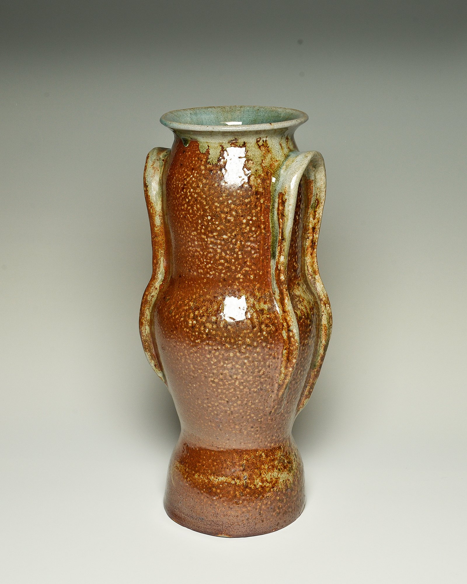 A ceramic vase with a textured surface and a wide mouth, featuring earthy tones of reddish-brown and muted green, set against a plain light background.