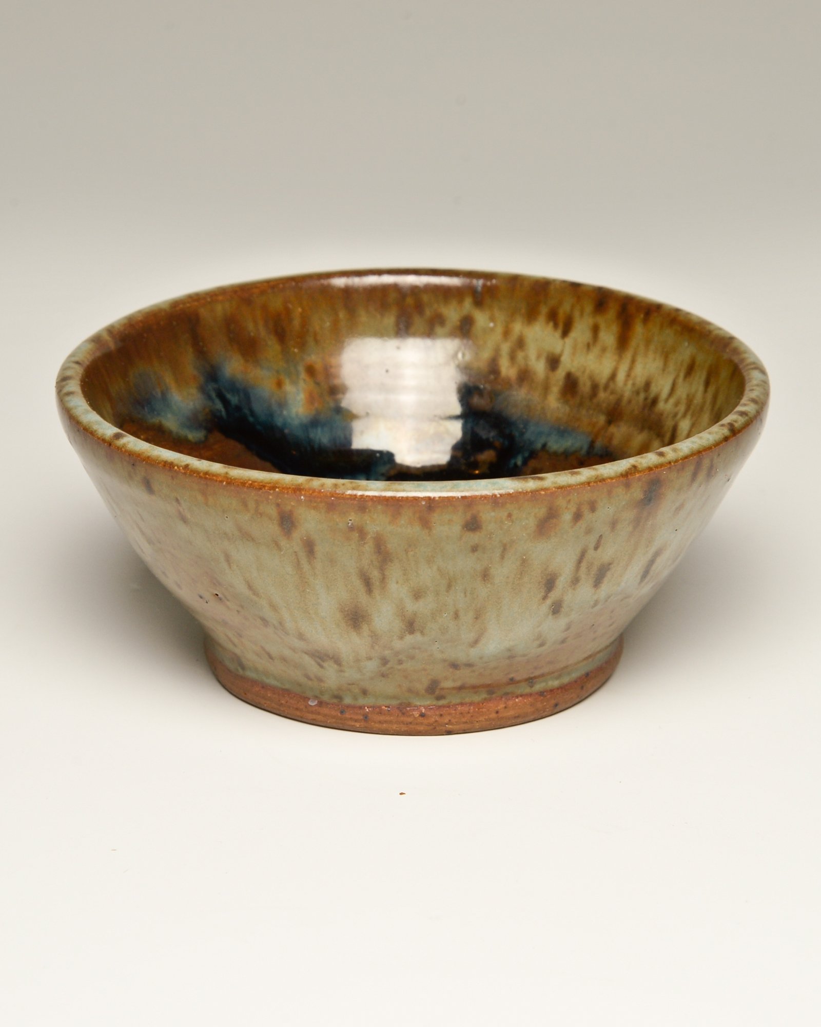 A ceramic bowl with a glazed, earthy brown and blue interior, and a rough, unglazed base, placed on a plain white surface.