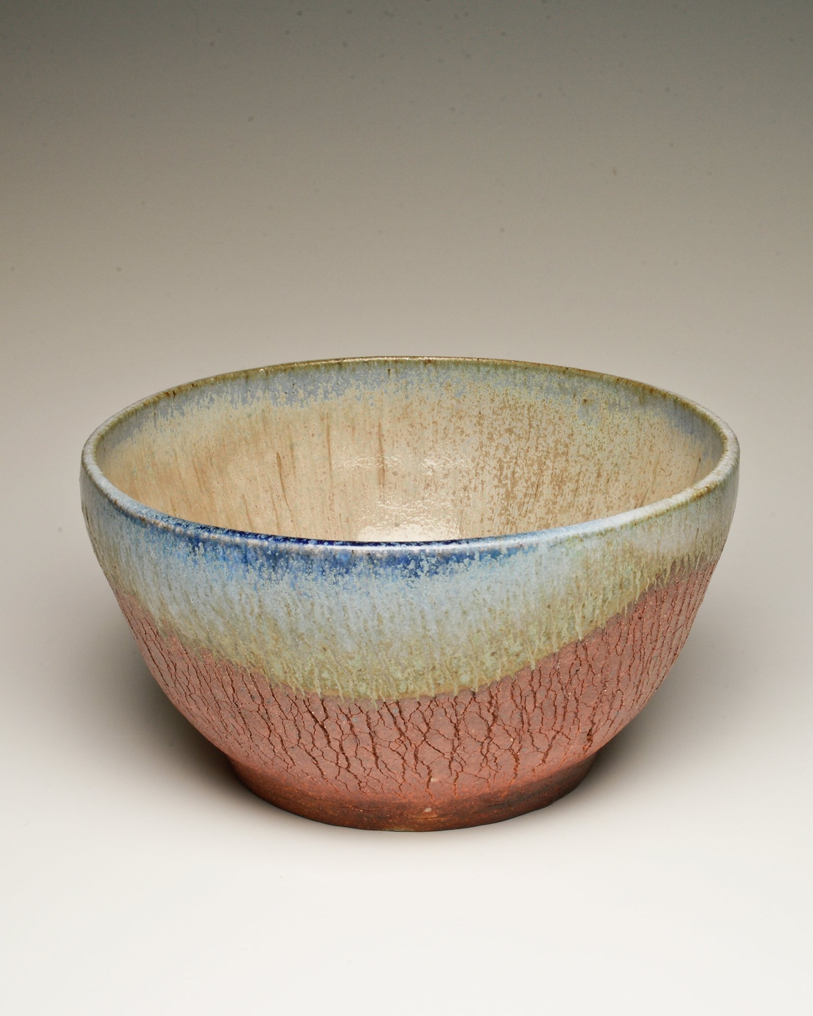 A handmade ceramic bowl with a crackled pinkish-red exterior and an interior with a gradient glaze transitioning from beige to blue.