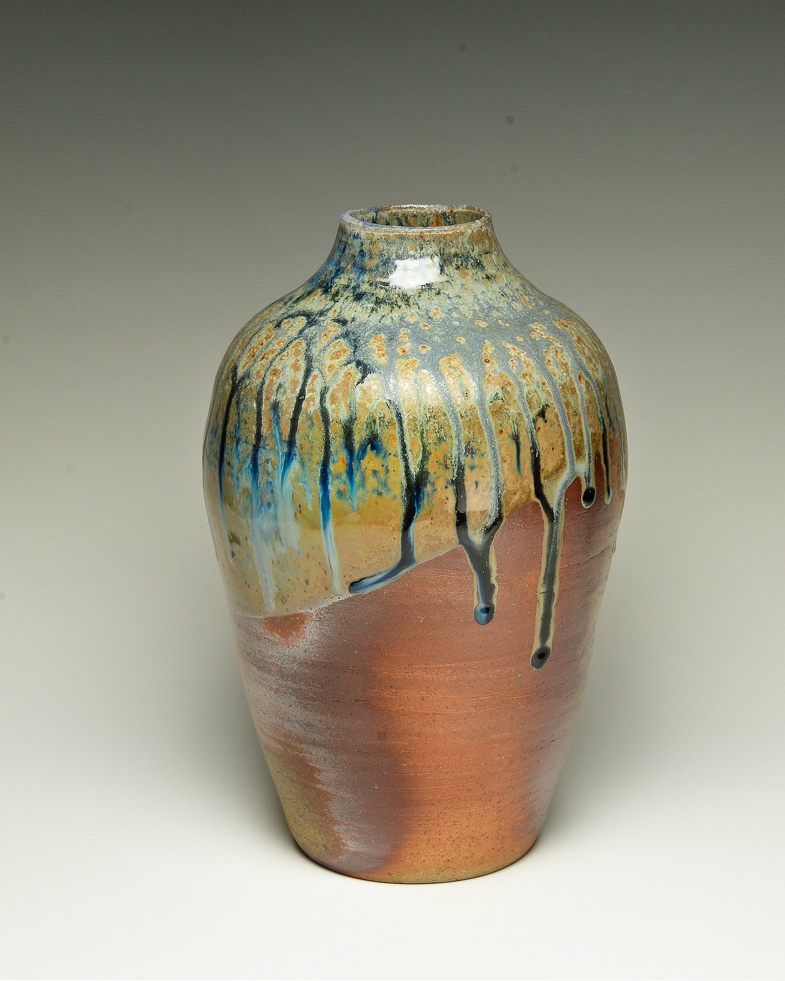 A ceramic vase with a multicolored drip glaze design, blending shades of blue, yellow, and black, on a white surface with a plain gray background.