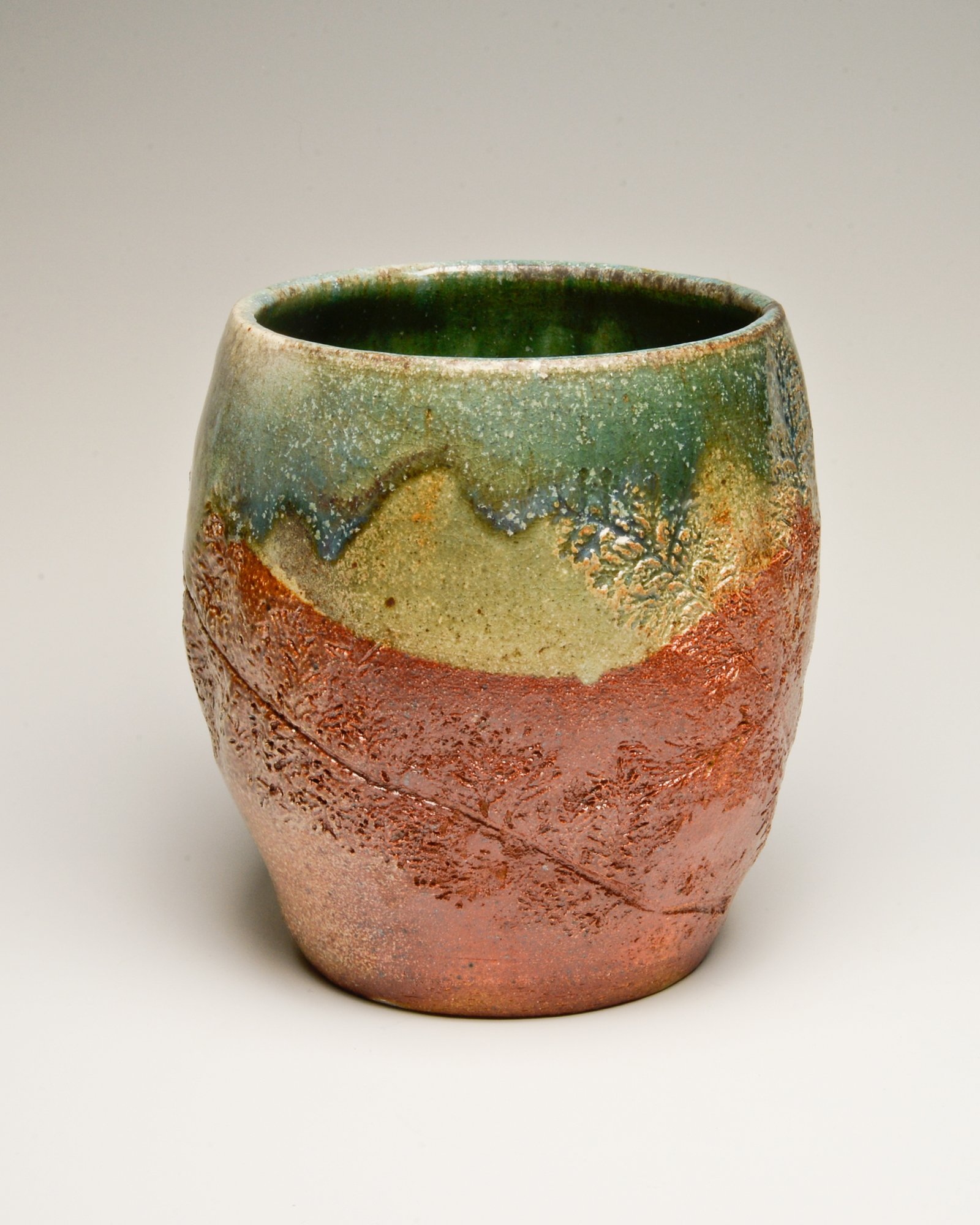 A ceramic vase with a textured surface and layered glaze in green, beige, and reddish-brown colors, set against a plain light background.