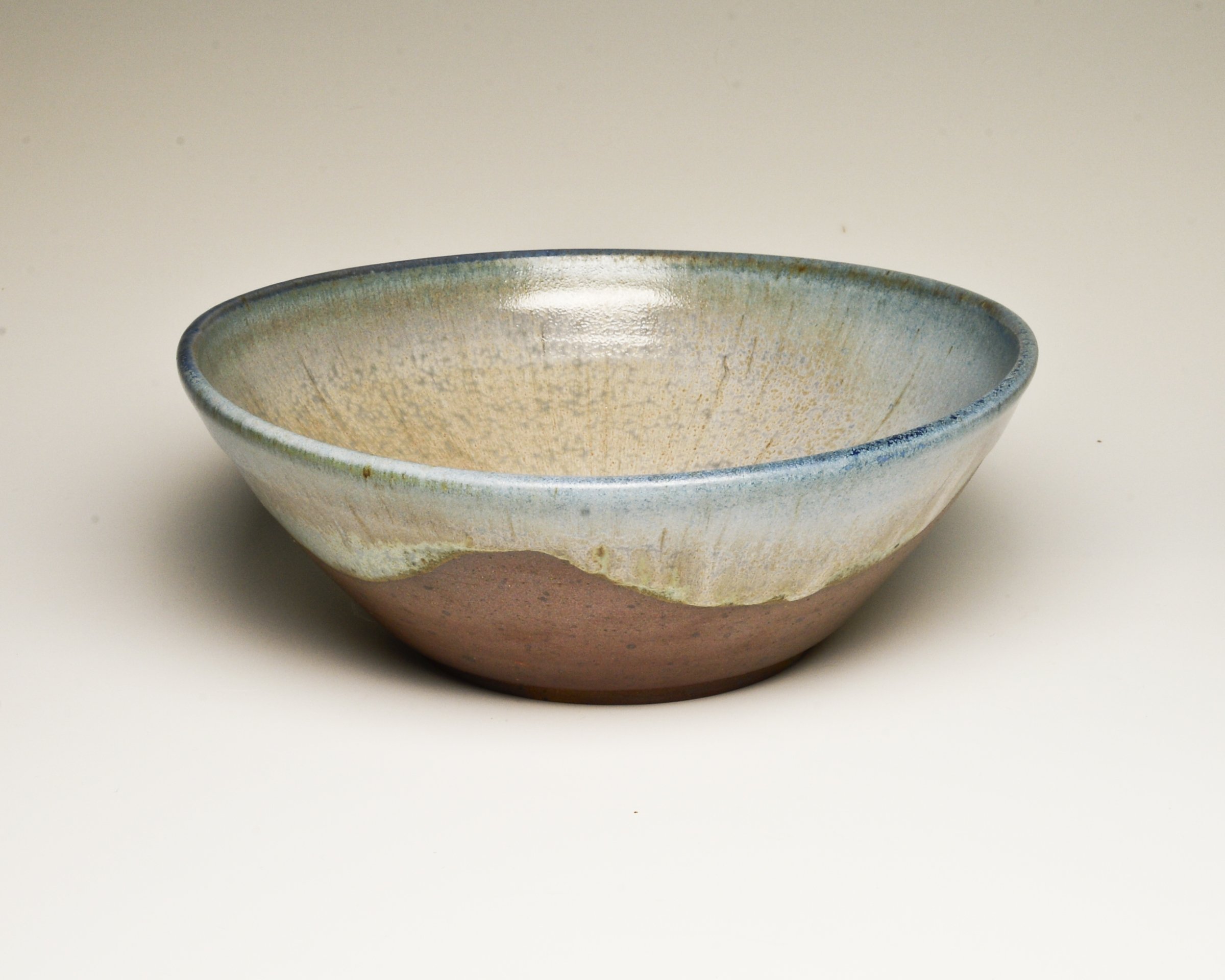 A ceramic bowl with a glazed interior that fades from light blue to beige, with a brown unglazed bottom, placed on a white surface.