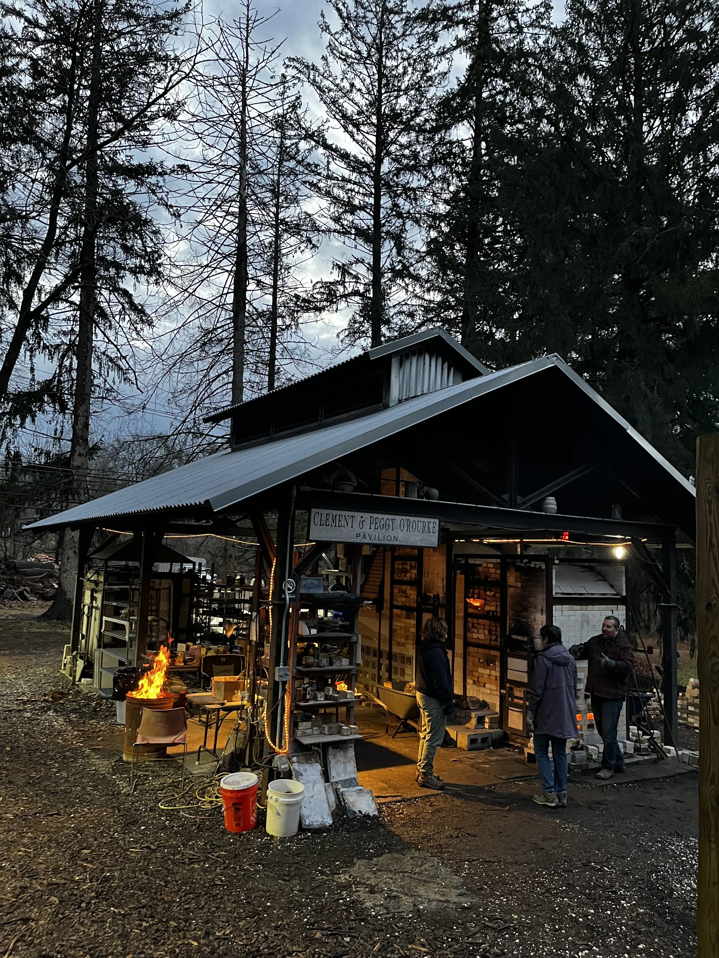 Outdoor gathering at a pavilion with people socializing, lit by string lights and a small fire, surrounded by trees in a wooded area during evening.