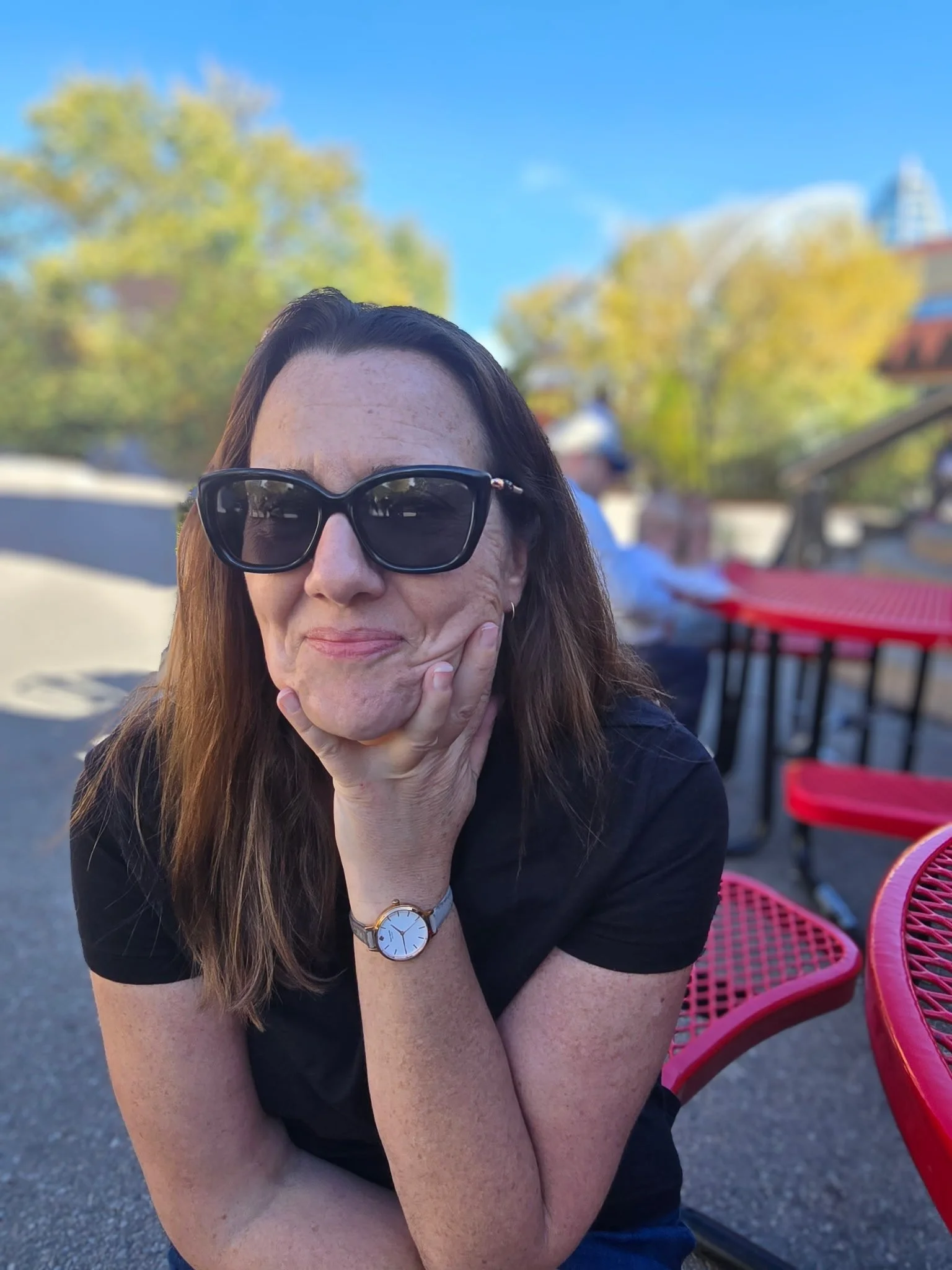 Close-up of a woman with long brown hair wearing sunglasses and a watch, sitting outdoors at a pink metal table with a red lattice chair, with trees and a blue sky in the background.