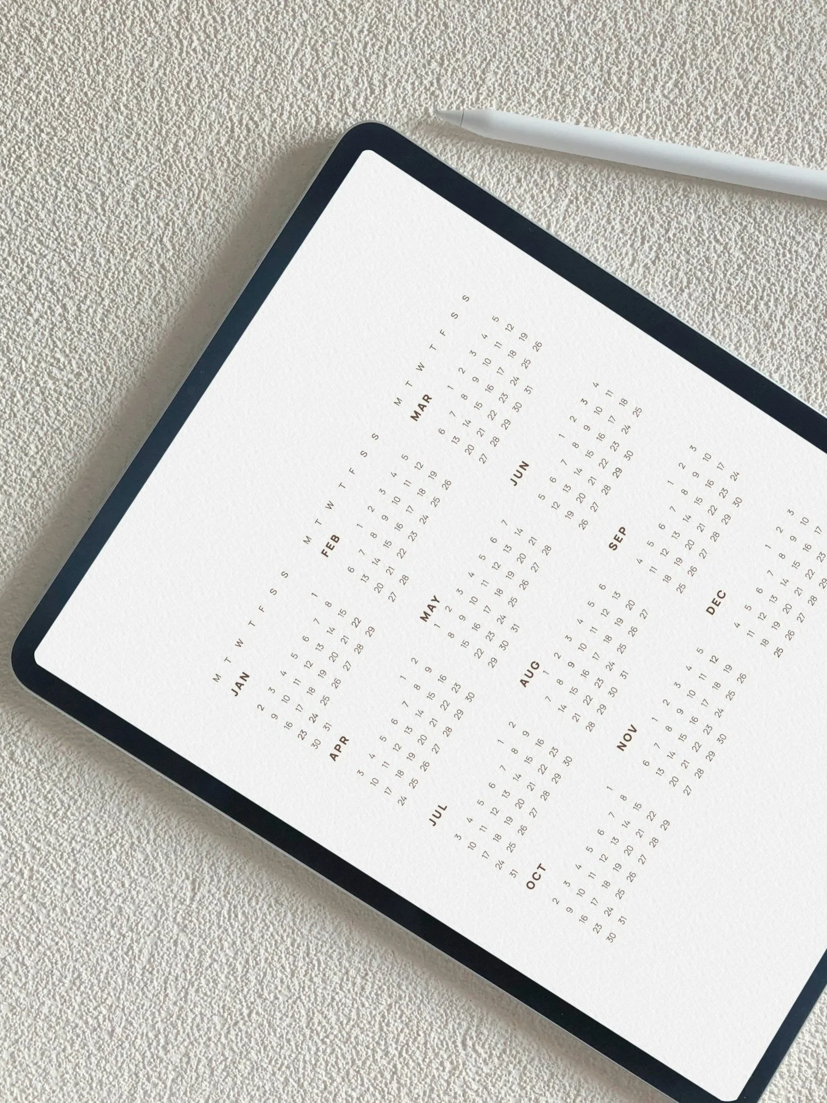 A digital tablet displaying a minimalist yearly calendar with months listed in columns, on a textured white surface with a white stylus beside it.