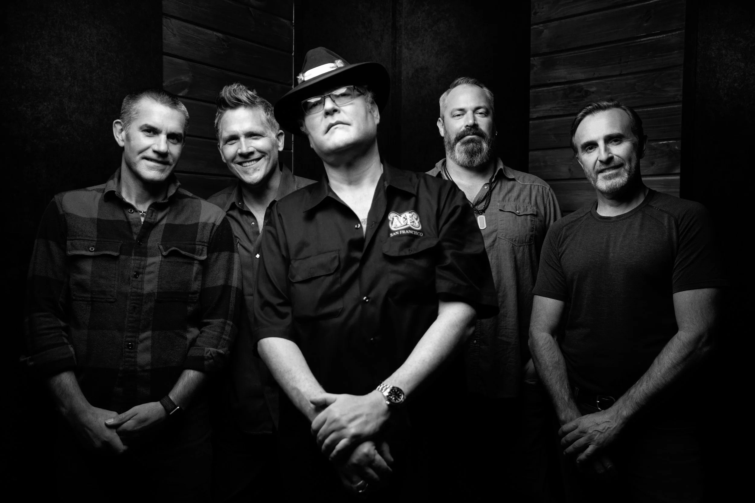 Black and white photo of five men standing in a row against a dark background with wooden paneling. The man in the center wears glasses, a hat, and a jacket with a logo, and stands with his hands clasped in front. The men have various hairstyles and facial hair, with serious and confident expressions.