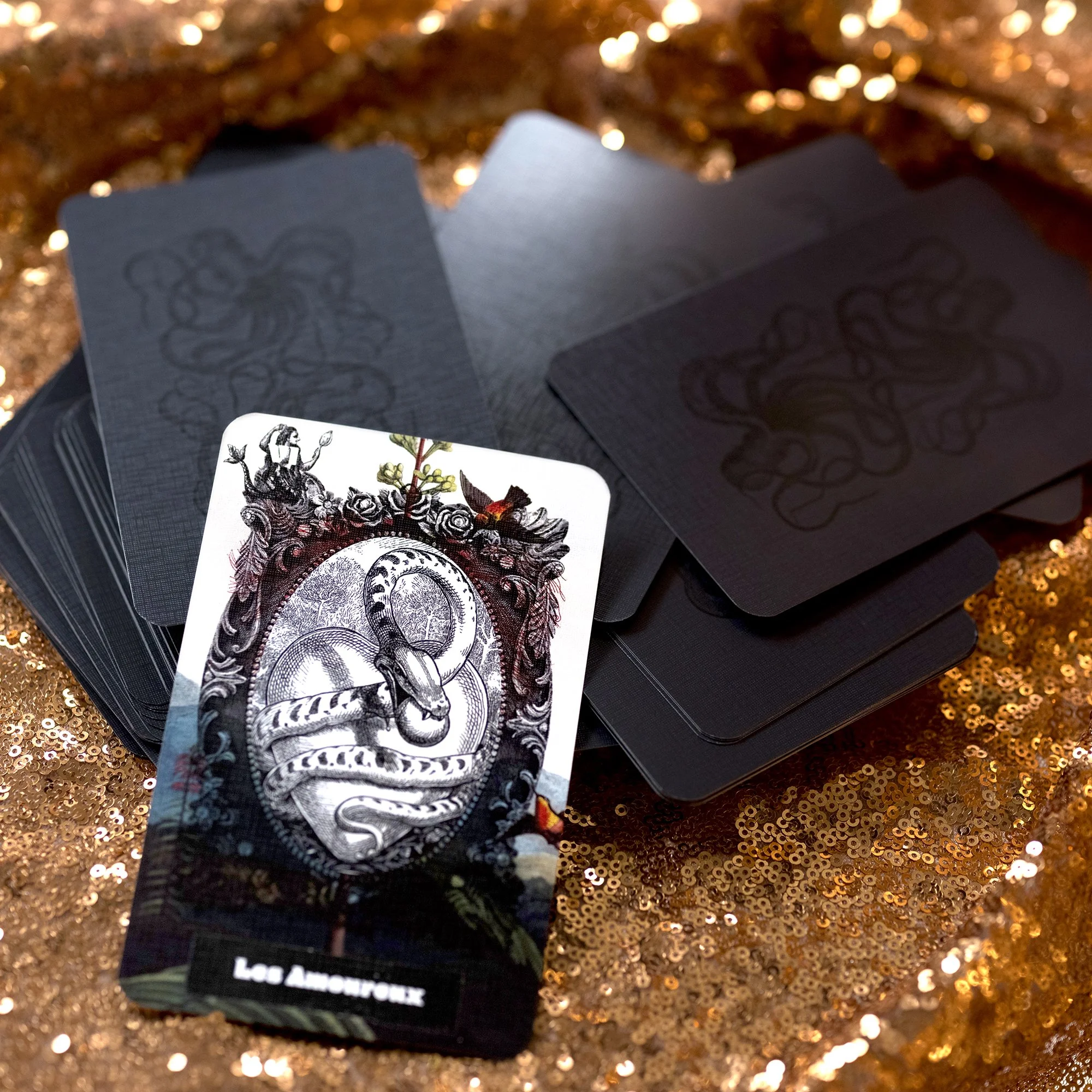 A deck of black tarot or playing cards with intricate white designs, placed on a gold sequin surface with some partially visible face-up cards showing detailed illustrations.