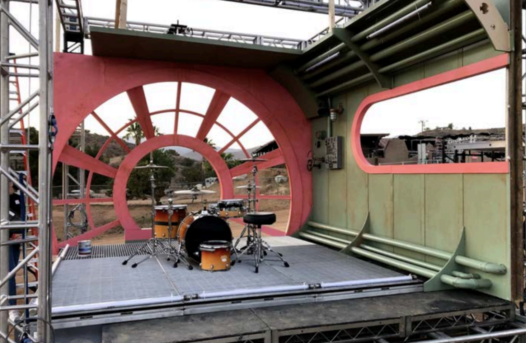A stage setup resembling a spaceship or futuristic environment with a pink and green color scheme, featuring a drum set and a stool, surrounded by metal scaffolding.
