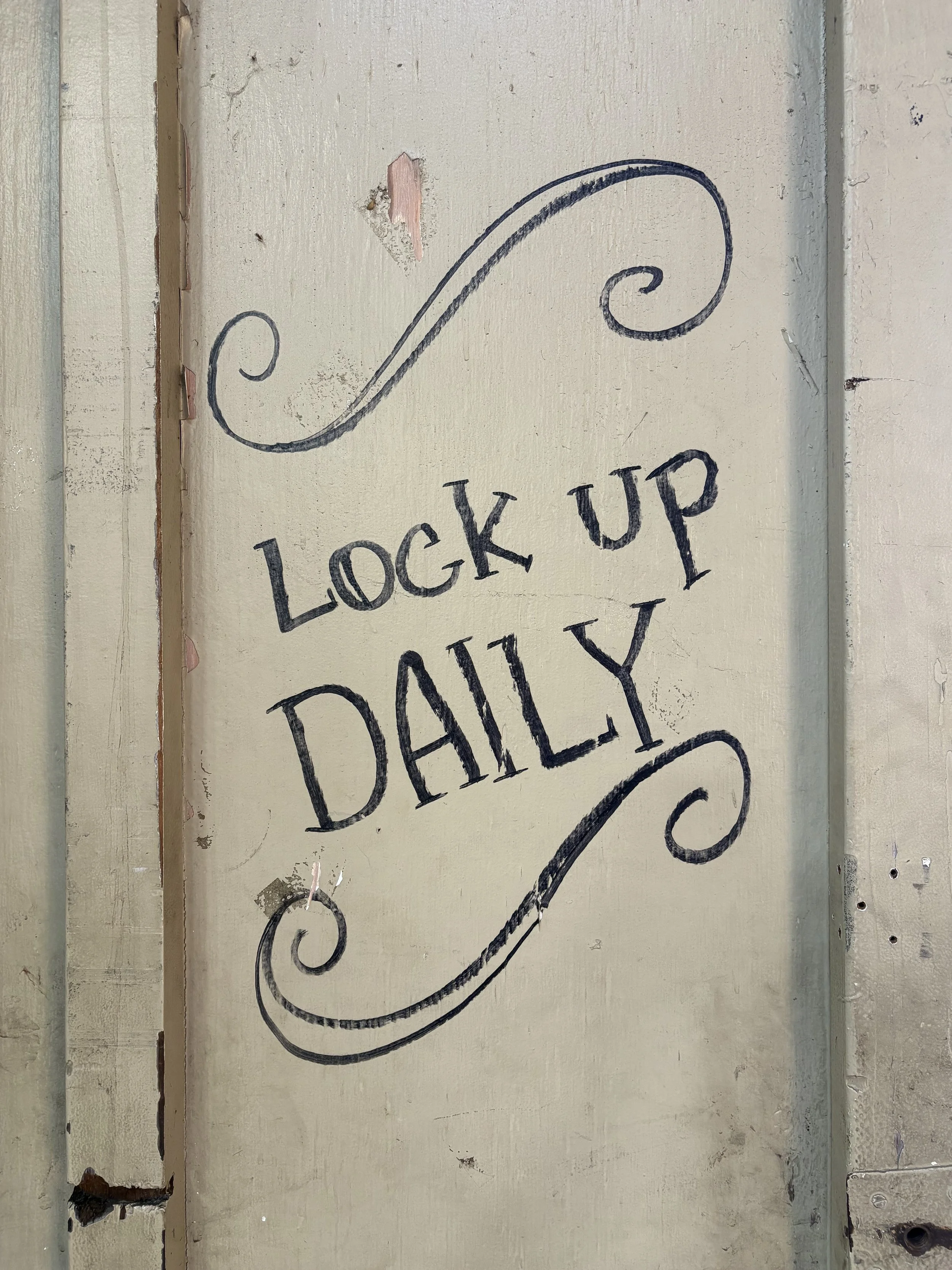 Hand-painted wooden sign reading 'Lock Up Daily' with decorative swirl design on a distressed door.