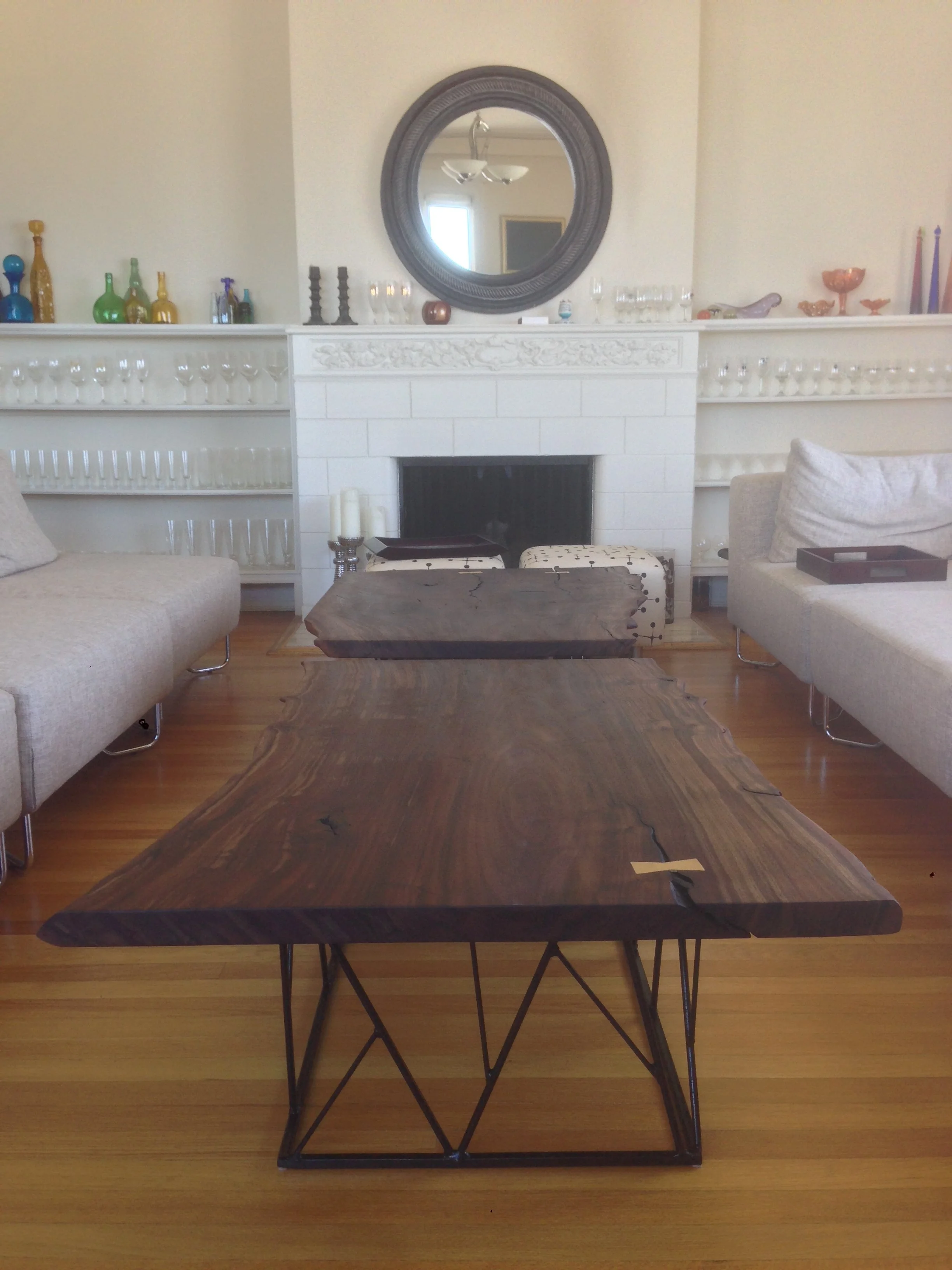 A living room with a dark wooden coffee table, two beige sofas, a white fireplace with decorative trim, a large round mirror, and a shelf with colorful glass bottles and decorative objects.