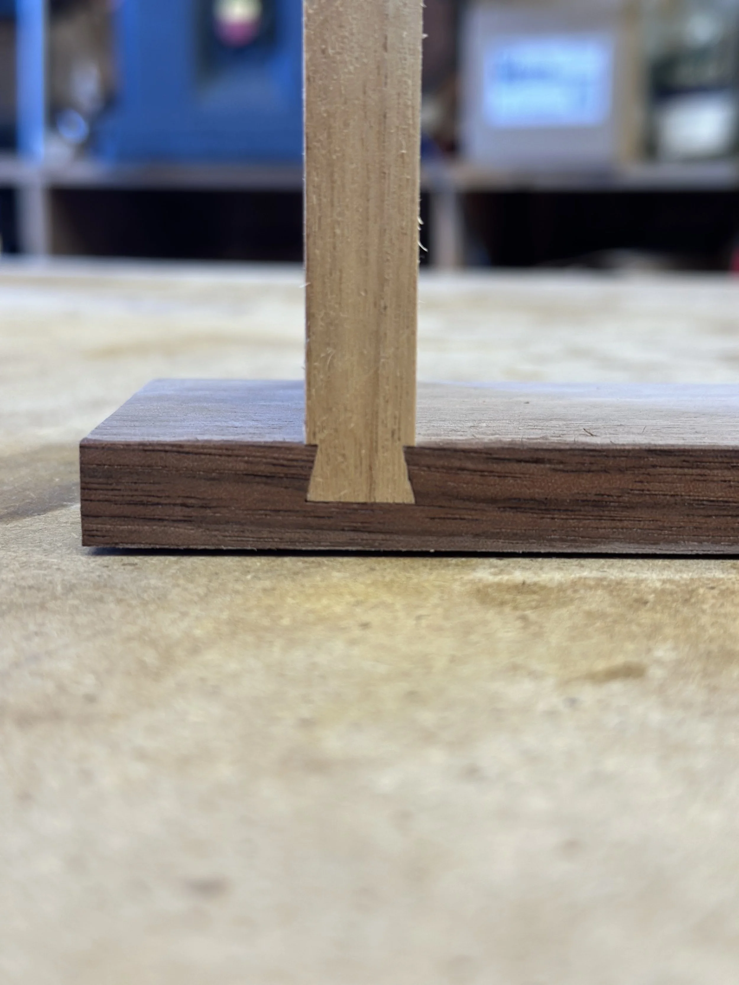 Close-up of a small wooden block with a wooden stick standing upright on it, placed on a work surface with a blurred workshop background.