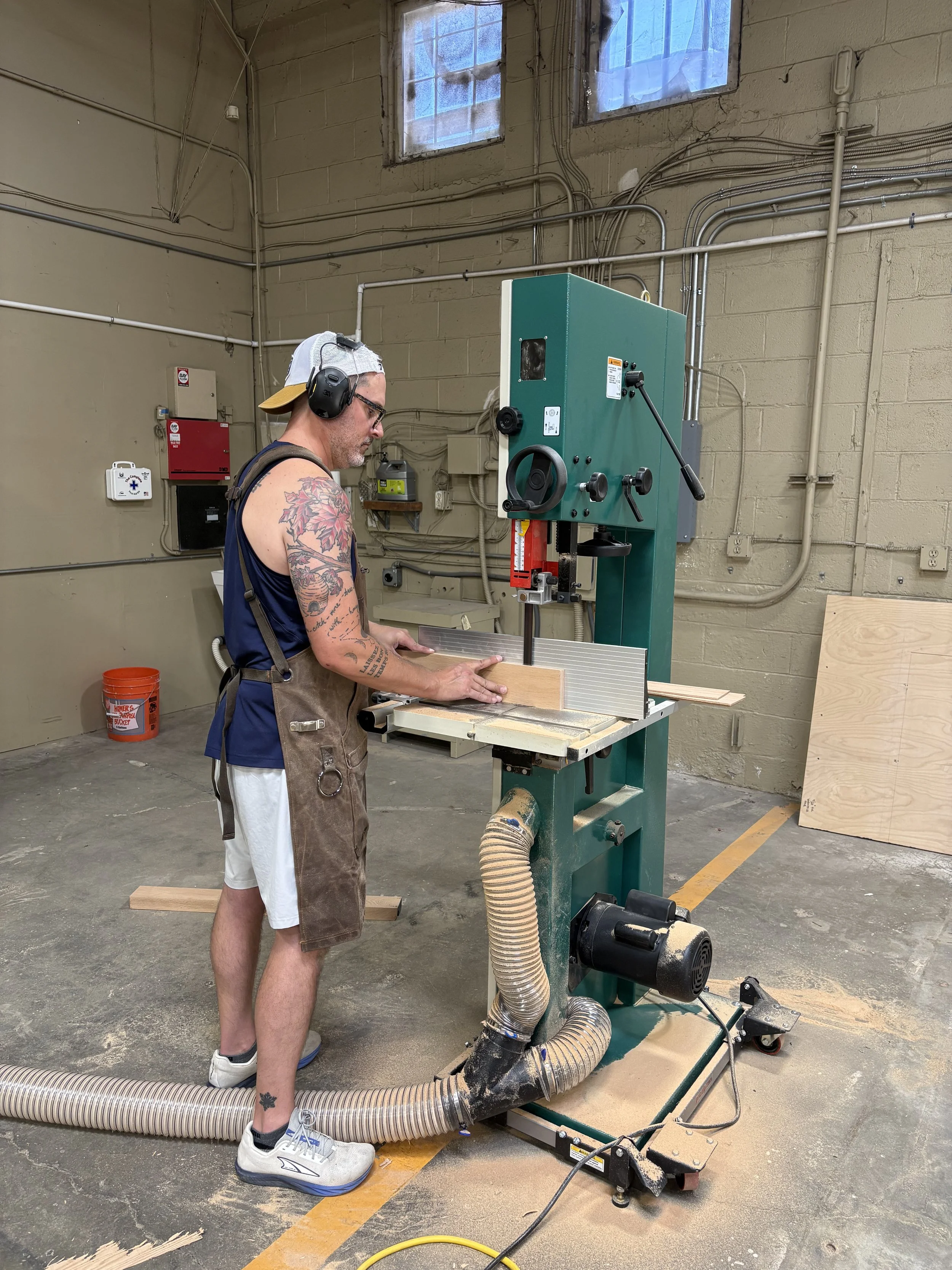 A man wearing ear protection and a backwards cap is using a bandsaw on a piece of wood in a workshop. He has tattoos on his arms and is wearing a sleeveless shirt and shorts. The workshop has electrical wires on the wall, a fire safety box, and a too
