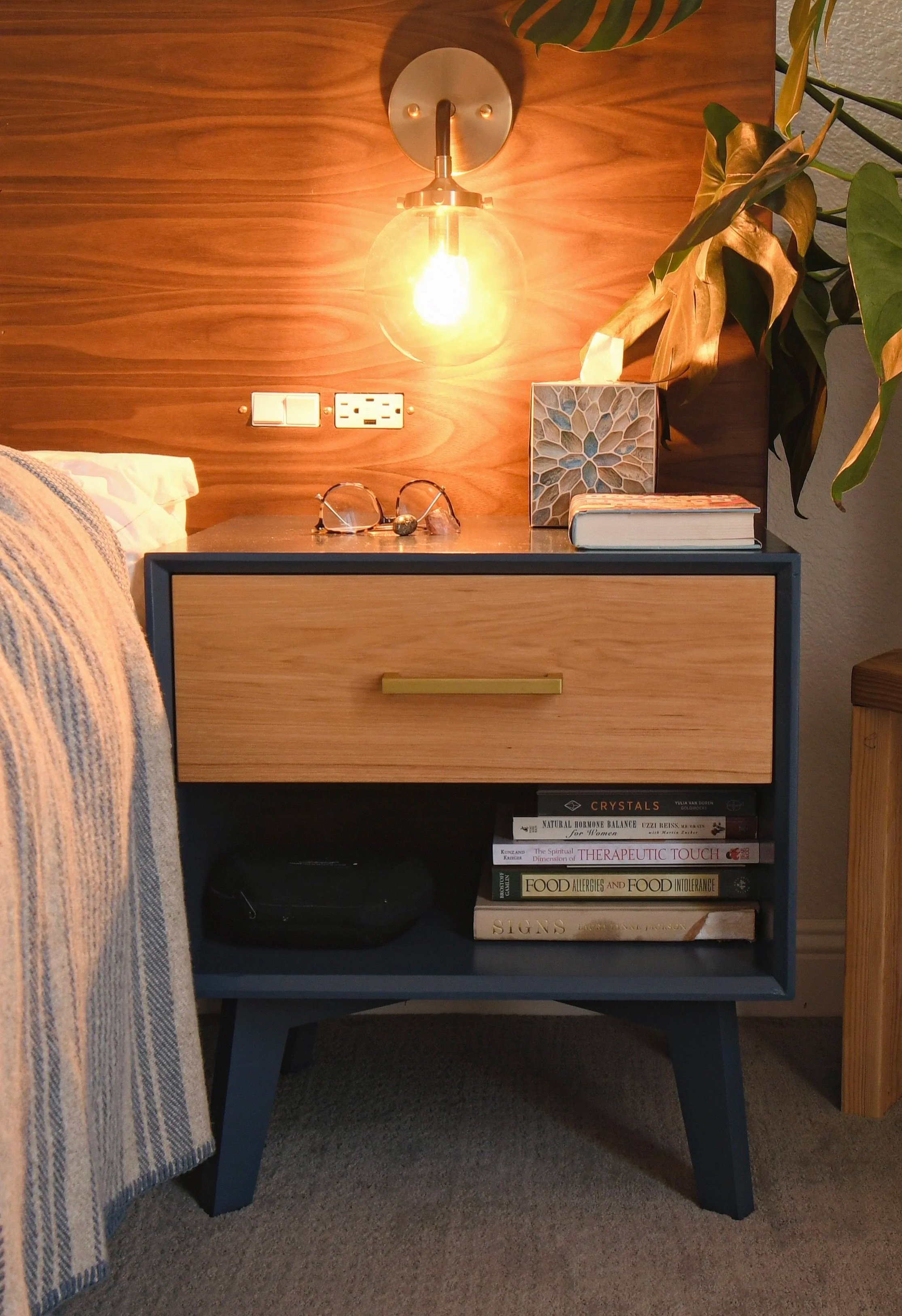 A modern bedside table with a blue drawer and black sides, placed on a carpeted floor. The table holds a pair of glasses, a decorative box, a book, and a tissue box. Above, a wooden headboard with a wall-mounted light fixture, electrical outlets, and