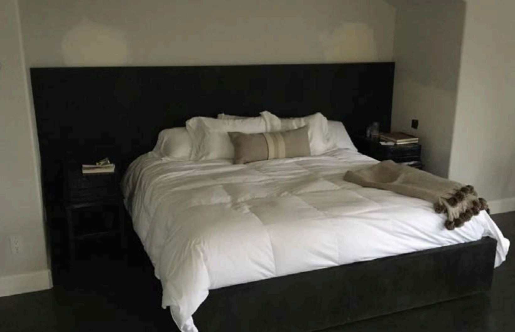 A neatly made bed with white bedding, a beige decorative pillow in the center, and a folded blanket with fringe at the foot of the bed. There are two dark nightstands on either side of the bed, each holding items like books and a water bottle. The he