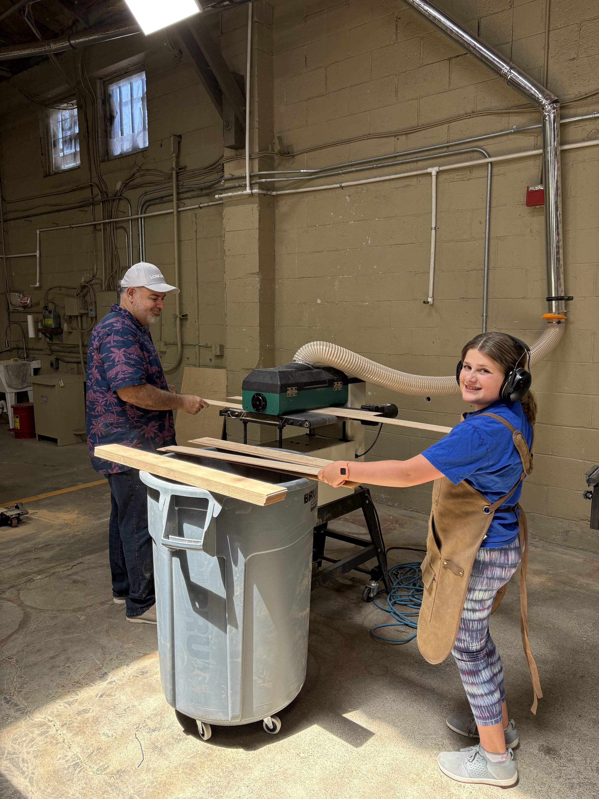 A man and a girl working in a woodworking workshop. The girl is using a machine that is connected to a large gray trash bin, and the man is working on a piece of wood nearby. The workshop has a concrete floor, a beige brick wall, and windows with sma