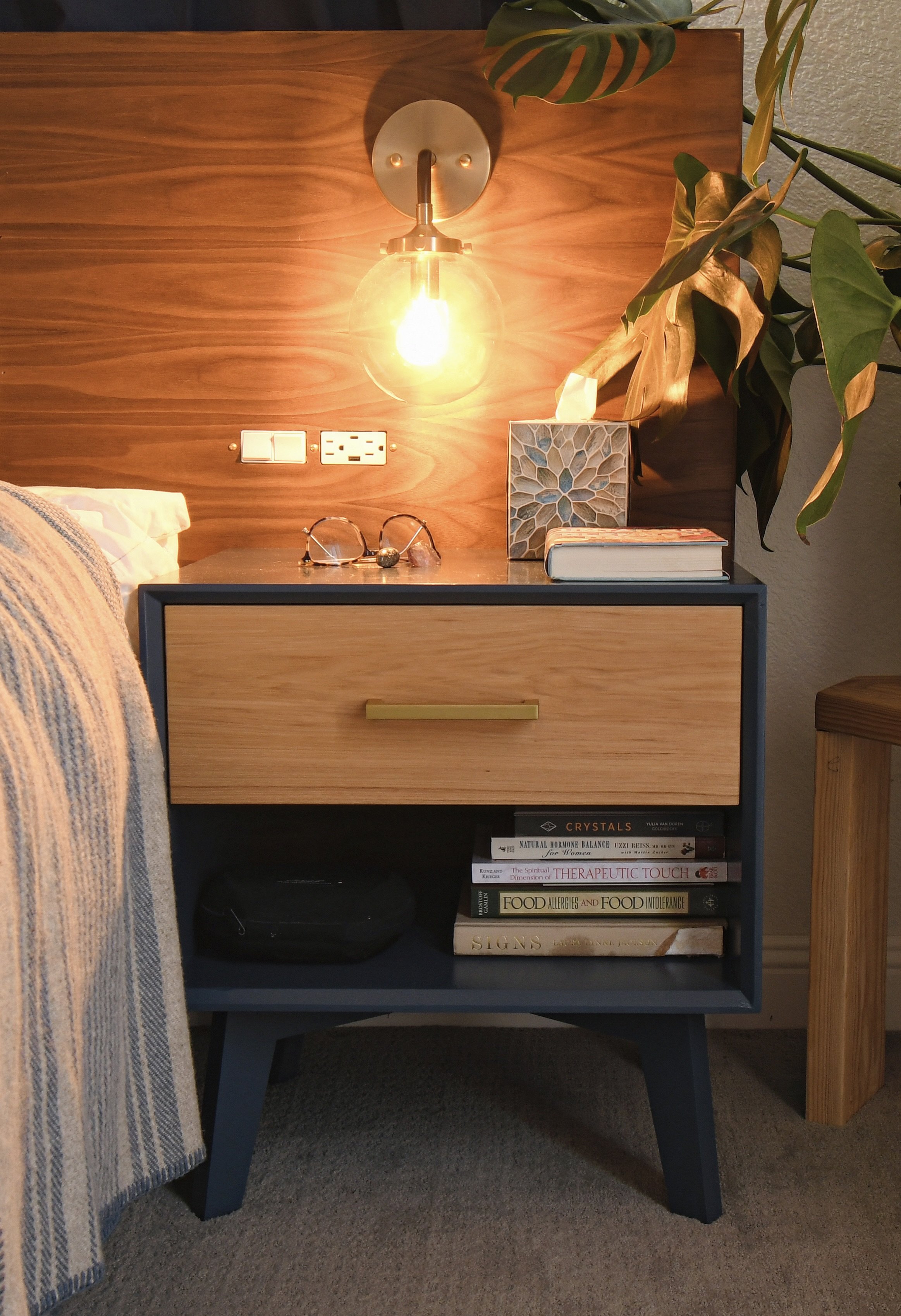 A modern nightstand with a wooden and dark blue finish, positioned against a wooden headboard and a textured wall. The nightstand holds a pair of glasses, a decorative box, a book, and a tissue box. The background includes a wall with electrical outl
