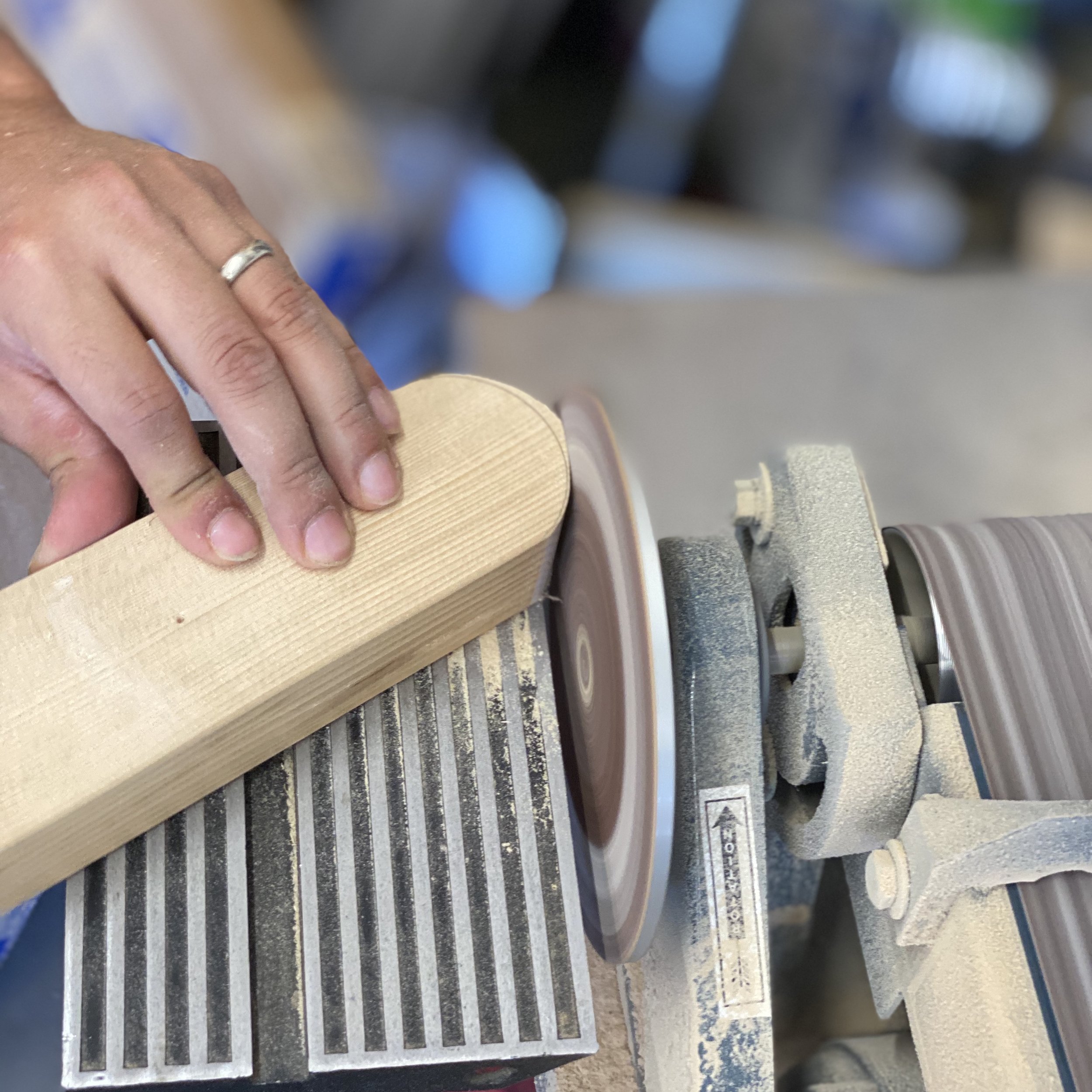 A person using a drum sander machine to smooth a circular piece of wood. The person's hand with a silver ring is guiding the wood on the sander, which is emitting some dust.
