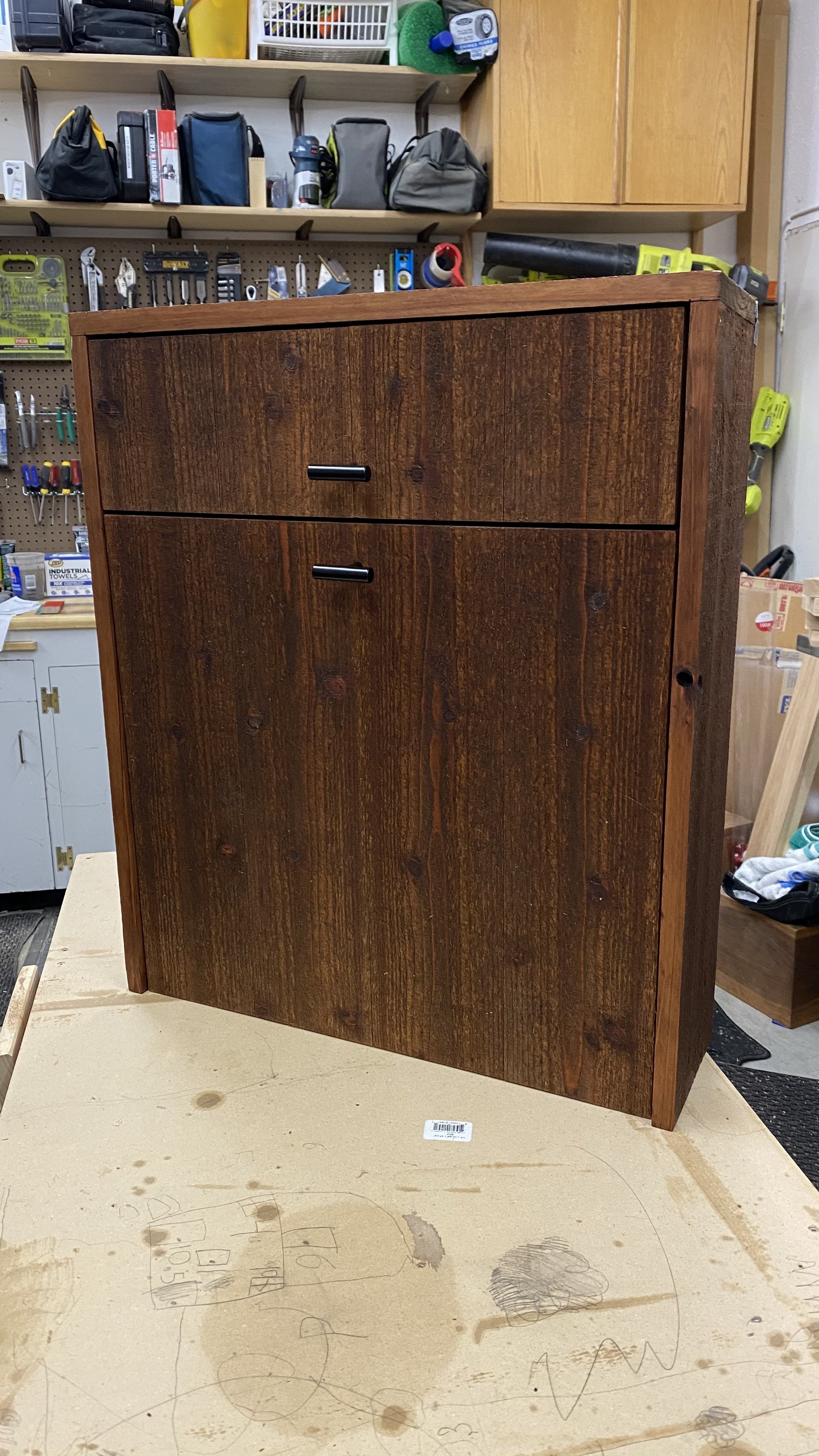 A wooden cabinet with two drawers is placed in a workshop or garage, surrounded by tools, boxes, and shelves.