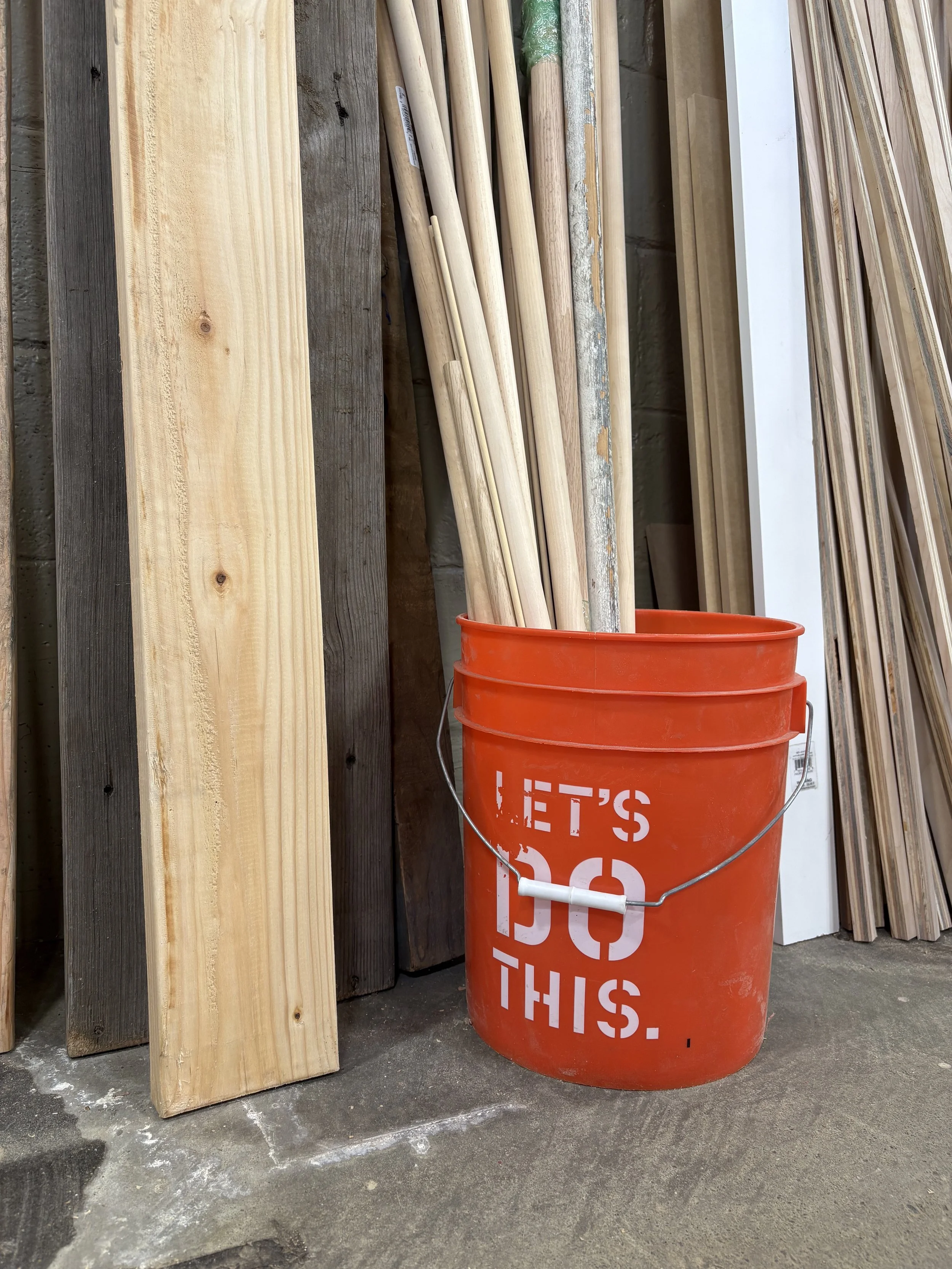 Red bucket with white text, containing wooden poles, surrounded by wood planks of different colors and sizes against a brick wall