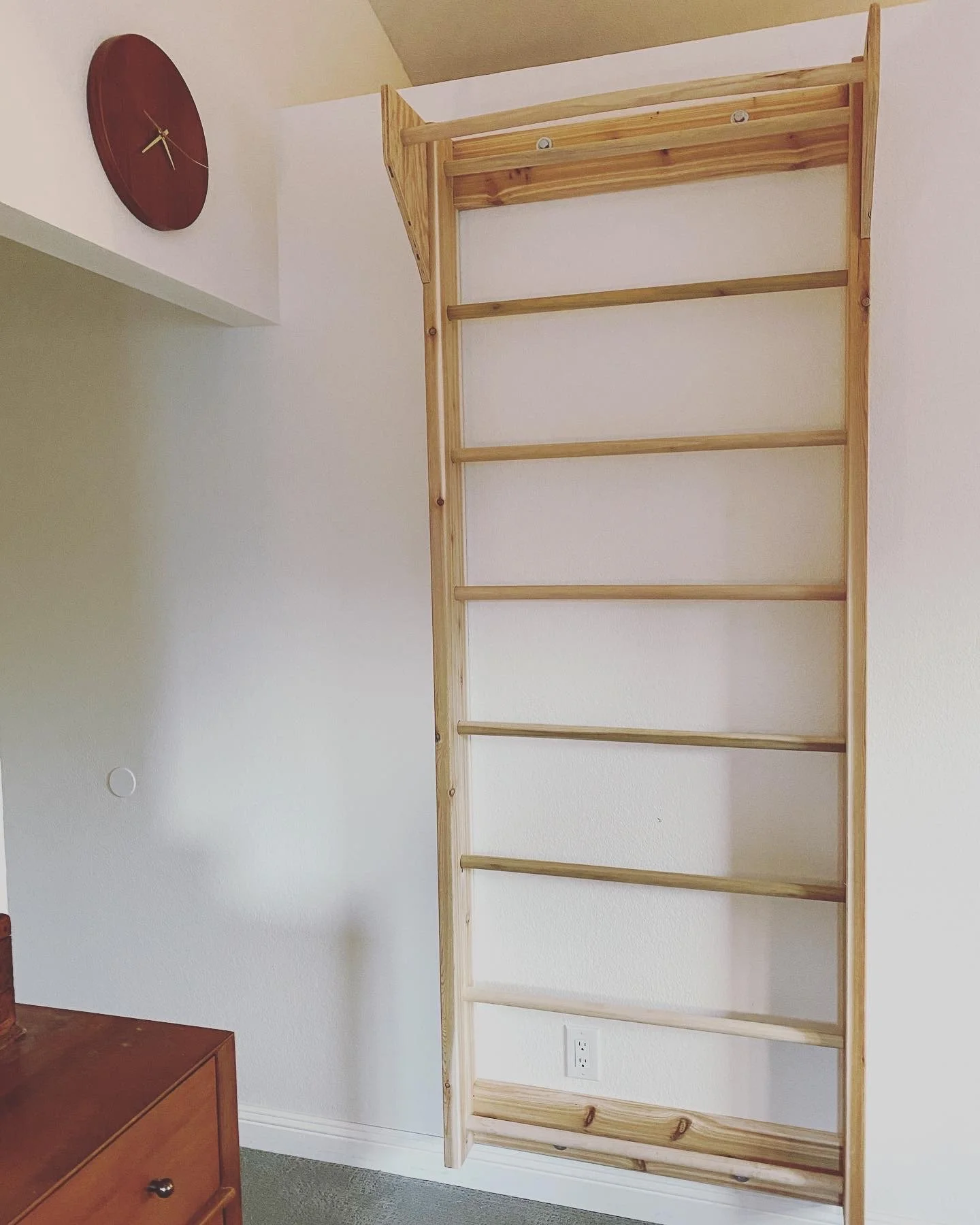 A wooden shelving unit with multiple empty shelves against a white wall, near a wall clock and a small wooden furniture piece in a room.