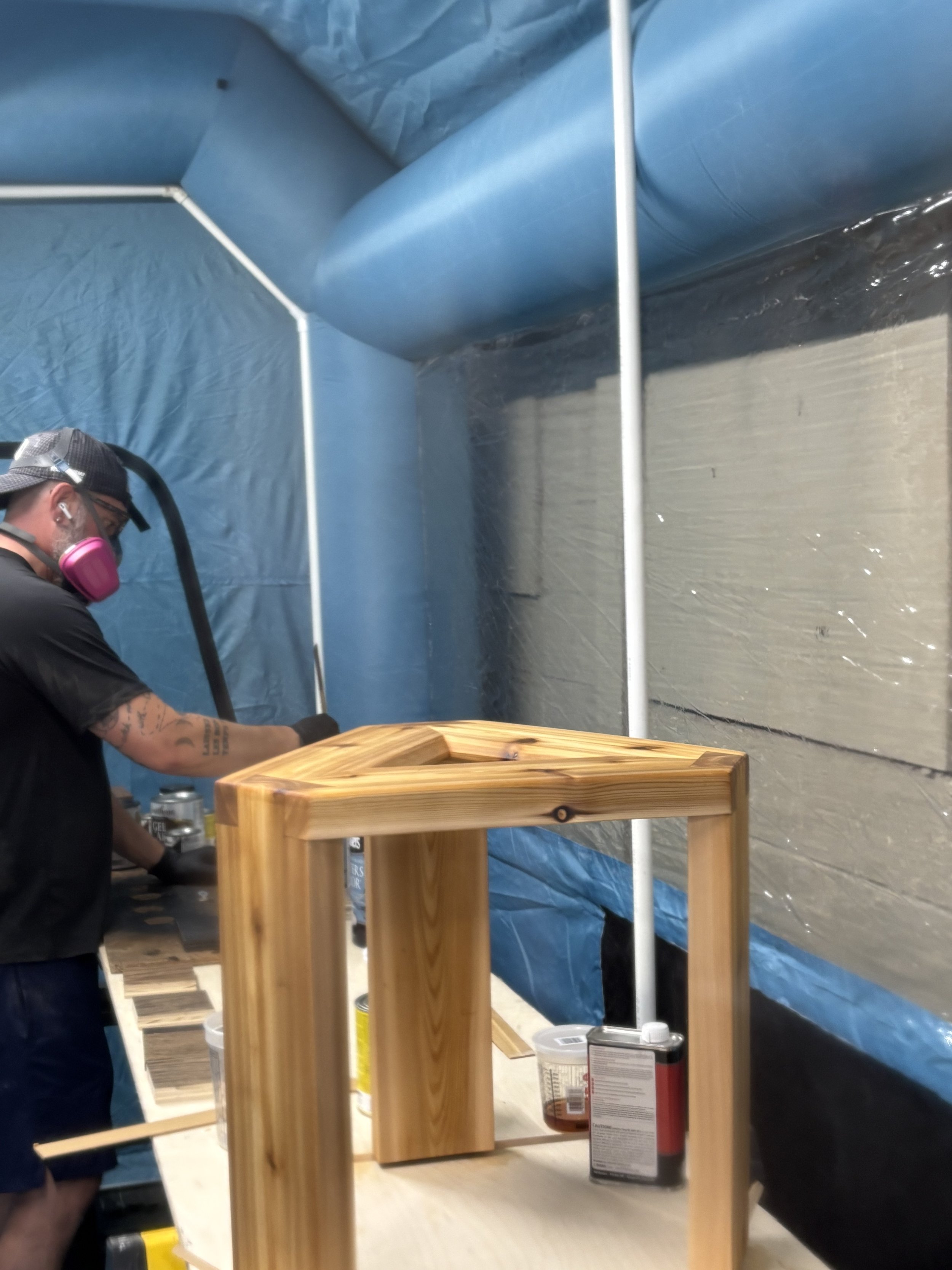 A person wearing goggles and a pink mask is sanding a wooden piece in a workshop with a blue plastic tarp covering the walls. There are cans of paint or varnish, a wooden furniture frame, and a small wooden table on the work surface.