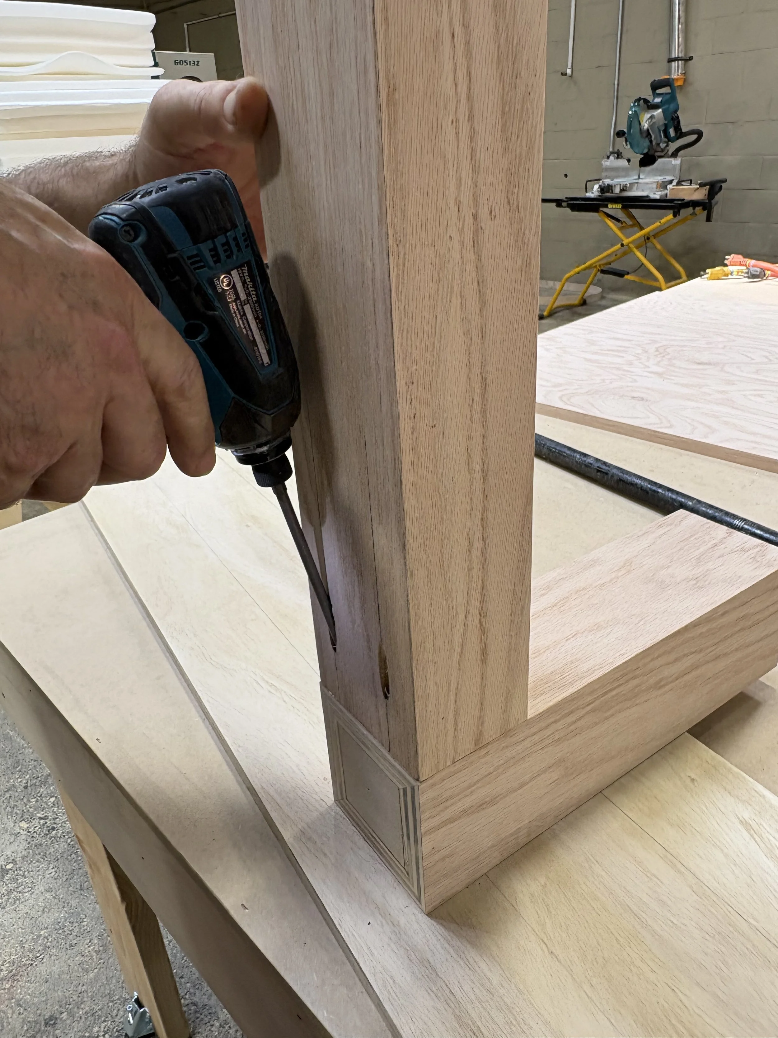 A person using a cordless drill to work on a wooden piece in a woodworking workshop, with saws and wood materials in the background.