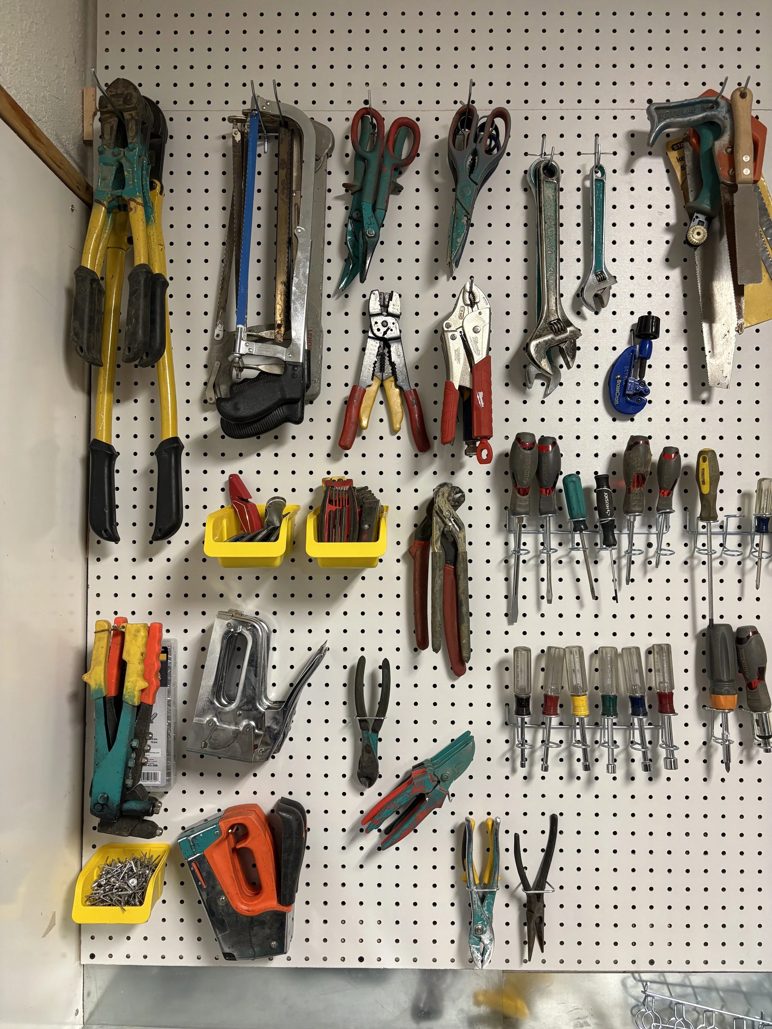 A pegboard mounted on a wall holds various hand tools, including scissors, wrenches, and screwdrivers. Two yellow containers at the bottom contain nails and screws.