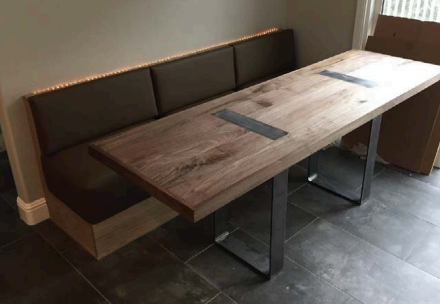 A wooden dining table with a natural finish and metal bases, positioned against a light-colored wall and near a window with curtains.