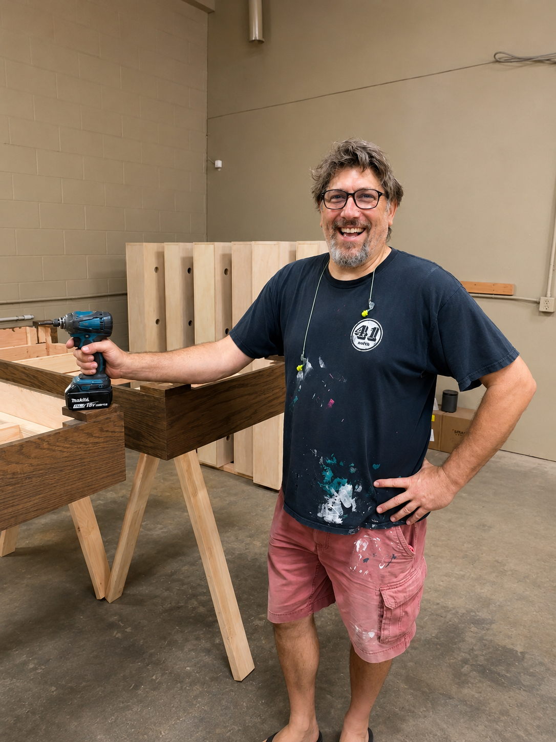 A man smiling and holding a cordless drill in a woodworking shop with wooden furniture and panels in the background.