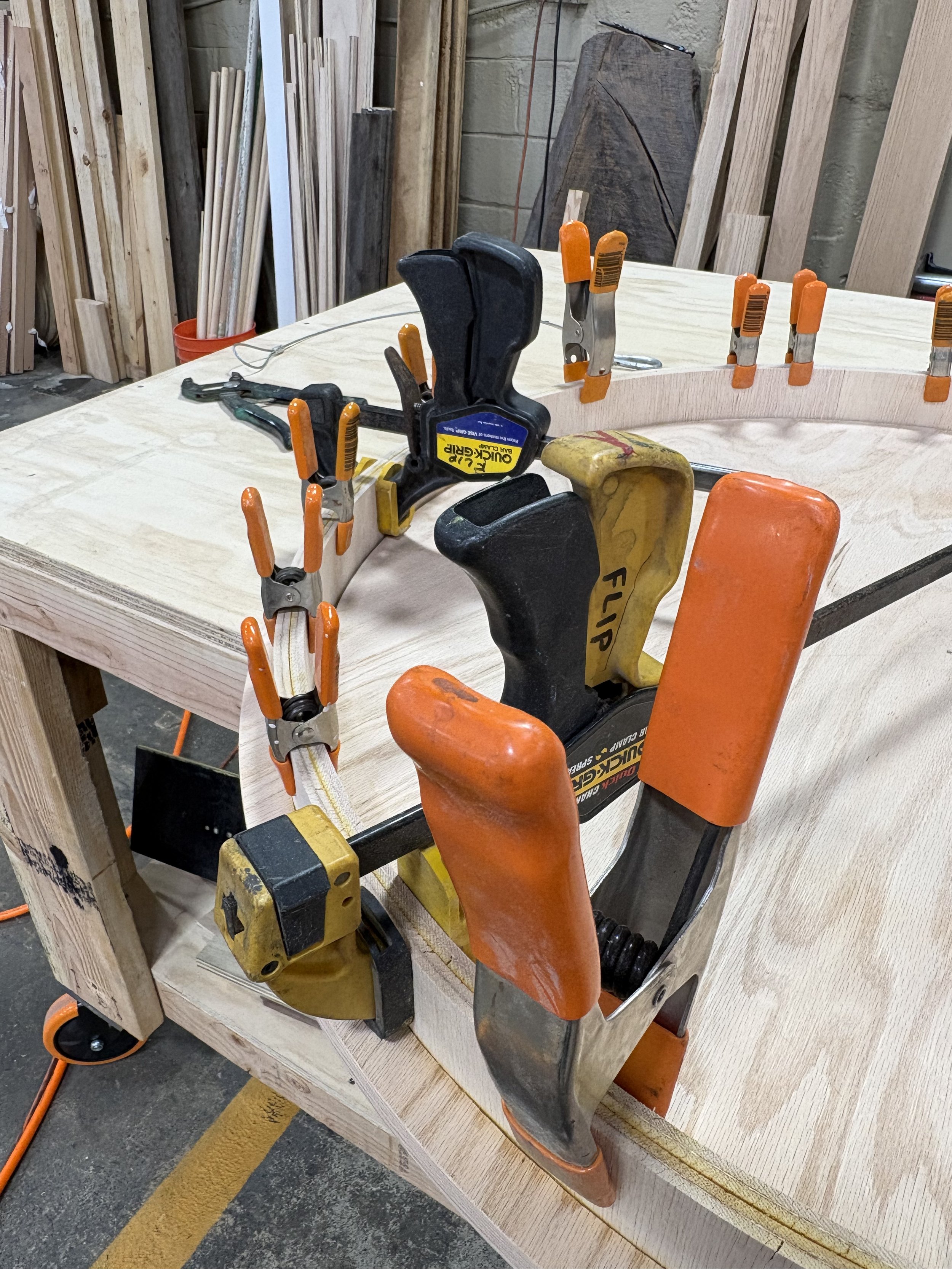 A woodworking project in progress with clamps securing a curved piece of wood to a work table, with various woodworking tools and materials in the background.